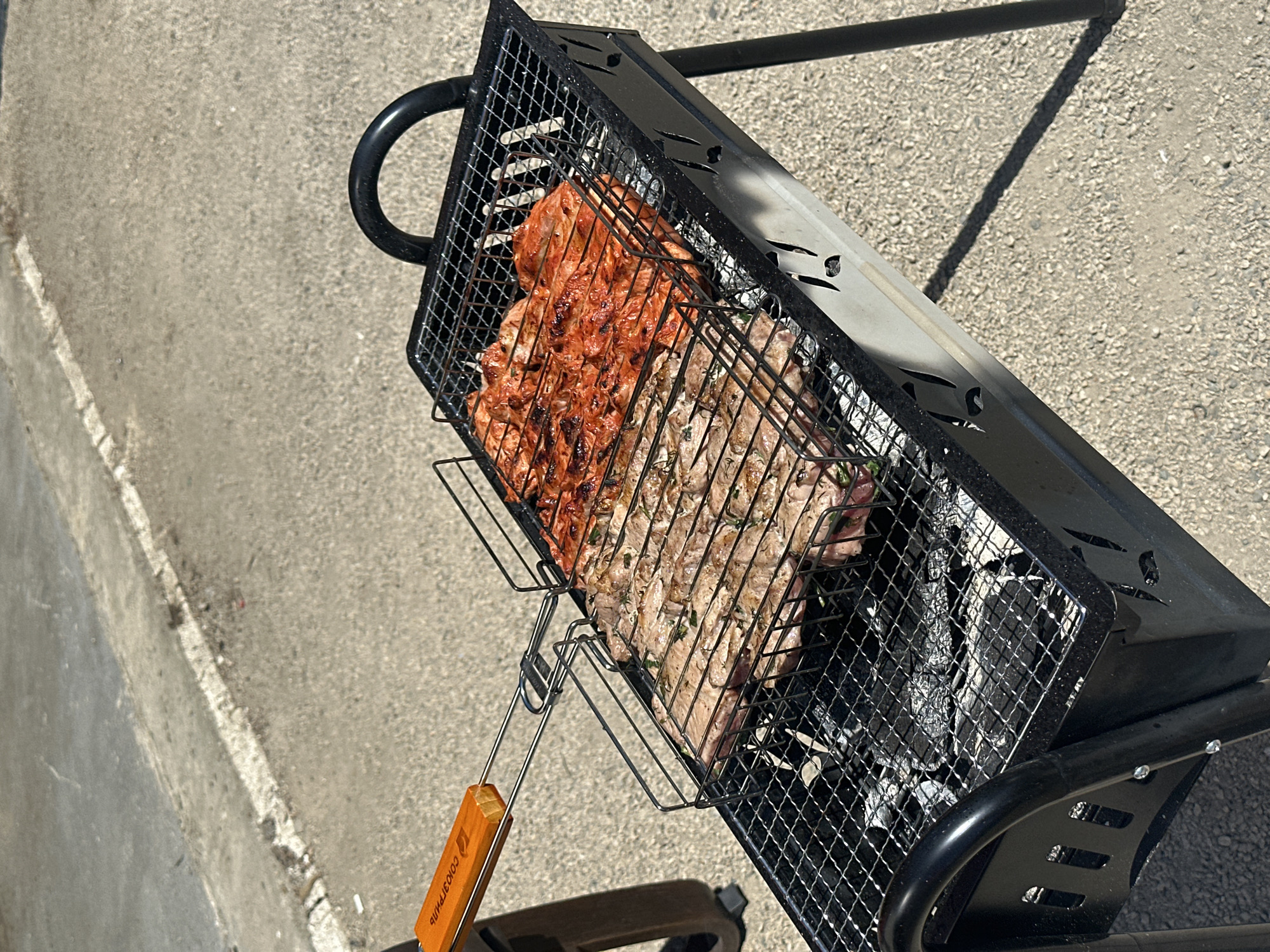 A medium-angle shot shows a black charcoal grill cooking meat outside. The grill is square and elevated on a wheeled frame. The upper wire grate contains two types of meat, one with a red marinade and the other with green herbs. Below the grate is charcoal. A long-handled, orange-handled tool is attached to the grill basket. The background is a gray pavement. The city of Blagoveshchensk, Russia is not determinable from this picture.