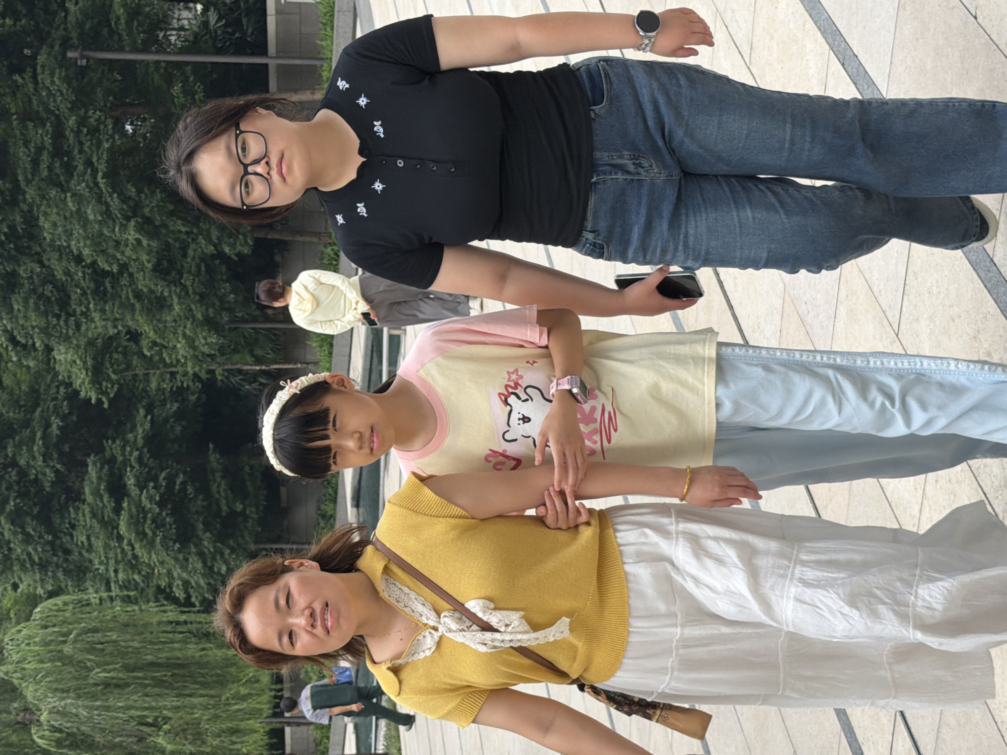 In Yuzhong District, China, three females walk on a paved outdoor area with trees and foliage in the background. A woman on the left, with medium brown hair, wears a yellow sleeveless top with a white lace collar and a white tiered skirt. She carries a brown crossbody bag. Next to her, a young female with black hair and a white headband wears a light yellow t-shirt with graphics and light blue pants. On the right, a young female with dark hair and glasses wears a black t-shirt with white embroidery and blue jeans. She holds a black smartphone in her left hand and wears a silver watch on her right wrist. In the background, a person in a light yellow hooded jacket stands facing away from the camera. Another person in a green vest and blue pants walks away from the camera on the left side of the frame. The ground is paved with light-colored square tiles.