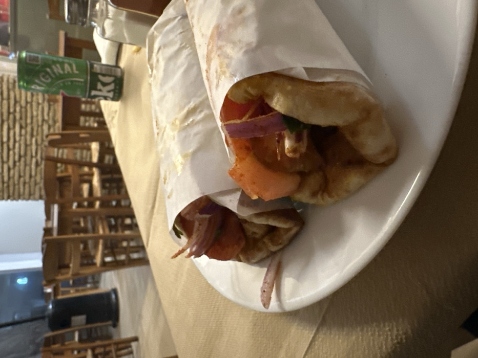 Two gyros or souvlaki wraps, encased in white paper, are the central focus of this indoor shot, resting on a white oval plate. The top wrap is partially open, revealing slices of red onion and tomato, along with pieces of meat. A similar wrap is visible underneath, with red onion peeking out. One long, thin piece of red onion has fallen onto the plate.
To the left of the plate, a green beverage can stands on a beige tablecloth, partially obscured. The word "ORIGINAL" is clearly visible on the can, along with the letters "Ke," suggesting it is a KEO beer, common in the region.
The background, which appears rotated 90 degrees clockwise from its natural orientation, shows a casual restaurant or tavern setting. A warm-toned, perhaps stone or brick, wall is visible behind several wooden chairs and tables, stacked or arranged in a slightly disordered manner. Further in the background, a blurry indoor area with indistinct features and some light sources can be seen. The overall impression is of a relaxed meal taking place in a traditional Greek eatery in Nea Erithrea.
