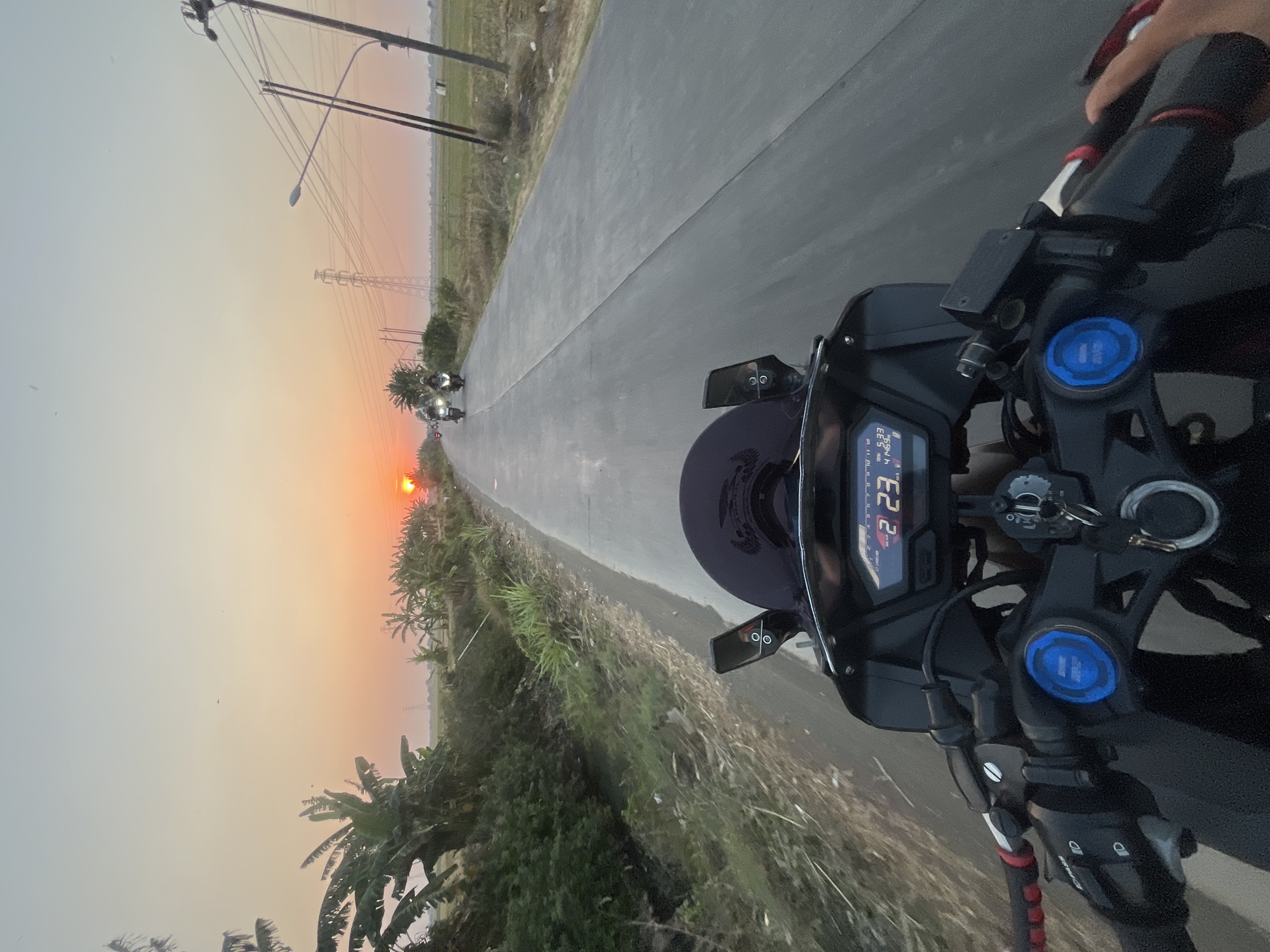 The image captures a first-person perspective of riding a motorcycle on a paved road at sunset, possibly in or near Batukali, Indonesia. The setting sun casts an orange glow across the horizon, illuminating a flat, rural landscape with fields and vegetation. Banana trees and other greenery line the left side of the road. In the distance, several other motorcycles are visible, following the same path. Power lines and utility poles are present along the right side of the road, indicating the presence of infrastructure. The motorcycle's dashboard shows a digital display with numbers, including "2 23" and "5:33". The handlebars and part of the motorcycle's front are in the foreground, with the rider's hand visible on the right grip. The overall scene depicts a peaceful, twilight journey through a rural Indonesian setting.