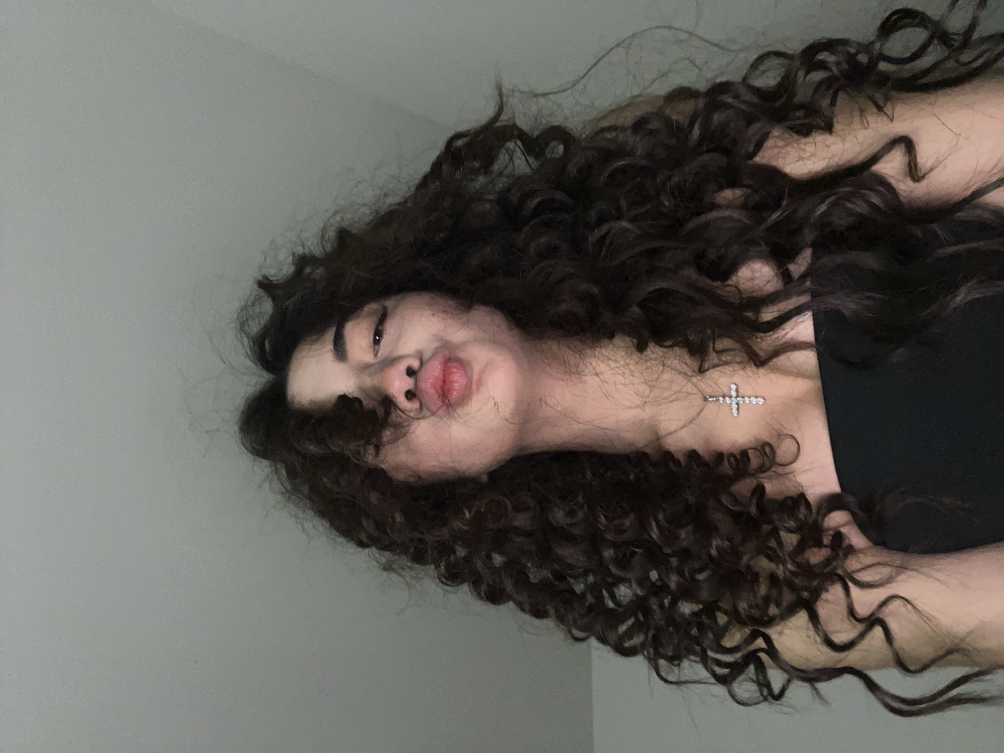 A young woman with long, dark, curly hair is looking up at the camera with her lips puckered. She is wearing a black top and a silver cross necklace. The background is a plain gray wall. There is no indication of the location being Chotěšov, Czechia.