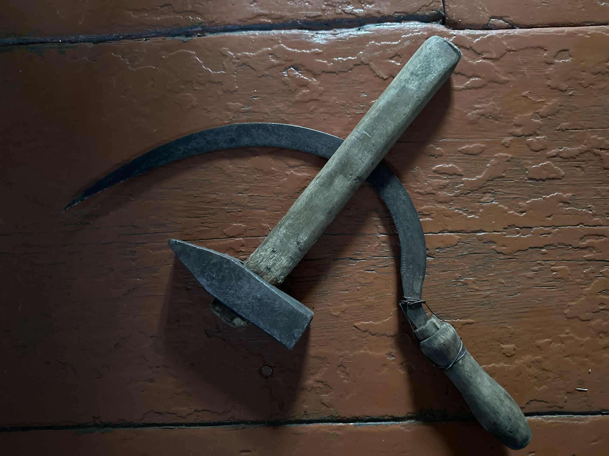 The image displays a hammer and sickle, iconic Soviet symbols, resting on a weathered, reddish-brown wooden surface. The hammer is positioned diagonally with its metal head towards the top left, and the sickle is curved around it, its serrated edge facing inwards. The tools appear old and well-used, with visible wear on the wooden handles and metal components. The lighting casts shadows on the surface, highlighting the texture and imperfections of the wood. There is no indication of the location in Biysk, Russia, or any people present.