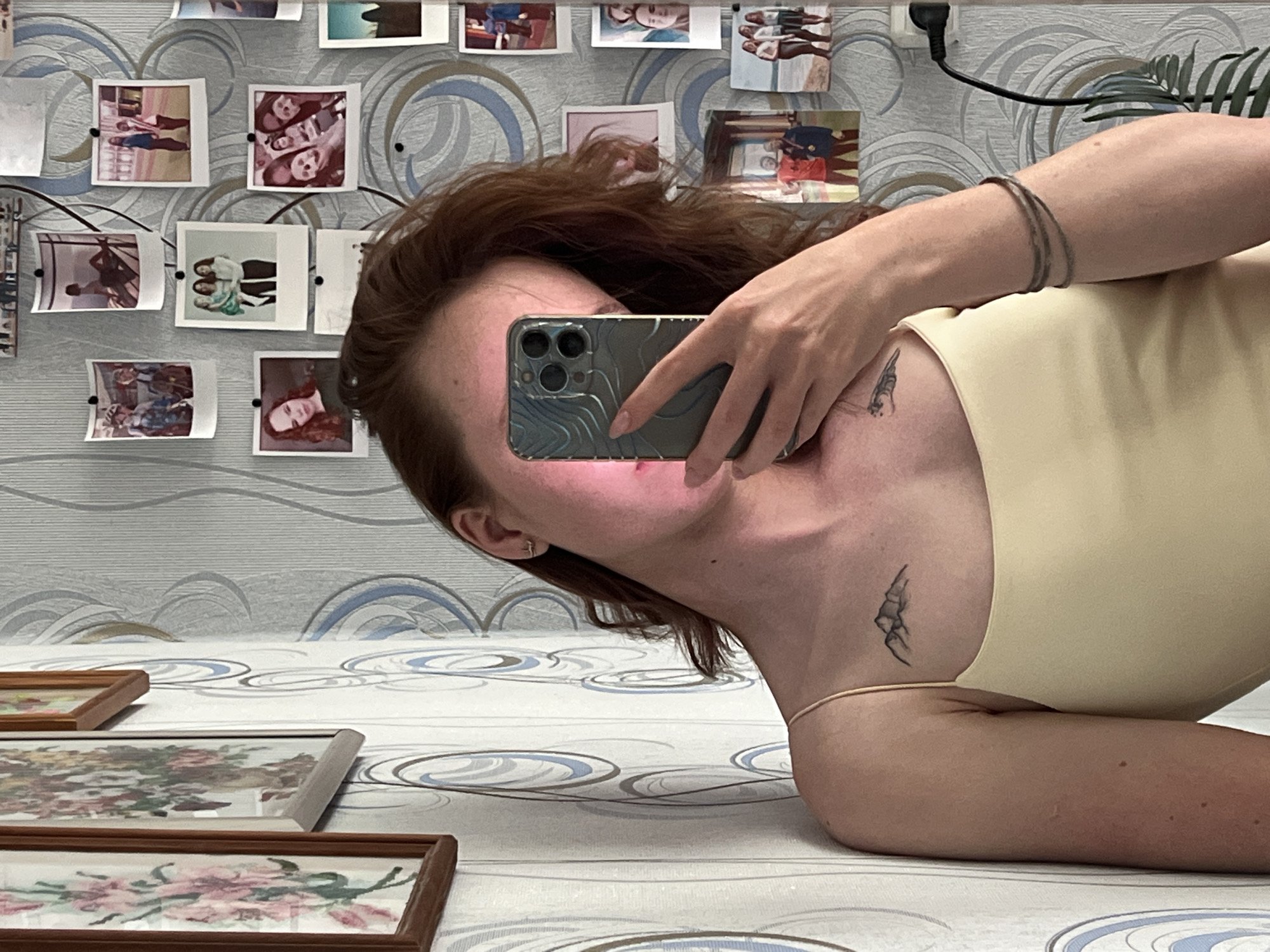 This image is a mirror selfie taken by a woman with reddish-brown hair, who is lying down or leaning back, causing her reflection and the background to appear inverted. Her face is largely obscured by the smartphone she holds, which has a multi-lens camera system and a textured, wave-like patterned case. She wears thin bracelets on her right wrist, a small stud earring in her left ear, and a light yellow top. A delicate black tattoo, resembling mountains or a landscape, is visible on her left shoulder/collarbone area.
The background, visible in the mirror, is a wall covered with a grey and light blue swirling patterned wallpaper. Numerous small, instant-print photographs are pinned to this wall, showcasing various people and scenes, suggesting personal memories or friends. In the upper right corner, an electrical outlet with a black cable plugged in, and a glimpse of a green plant are visible.
Below the mirrored reflection, in the foreground, the surface appears to be a bed or floor covered with a similar patterned fabric. Several wooden-framed pictures, likely embroidered or painted floral designs, are lying on this surface. The overall setting suggests a personal indoor space, possibly a bedroom, in Chaikovsky, Russia. No specific time of day or emotions are discernible, and no visible text is present.