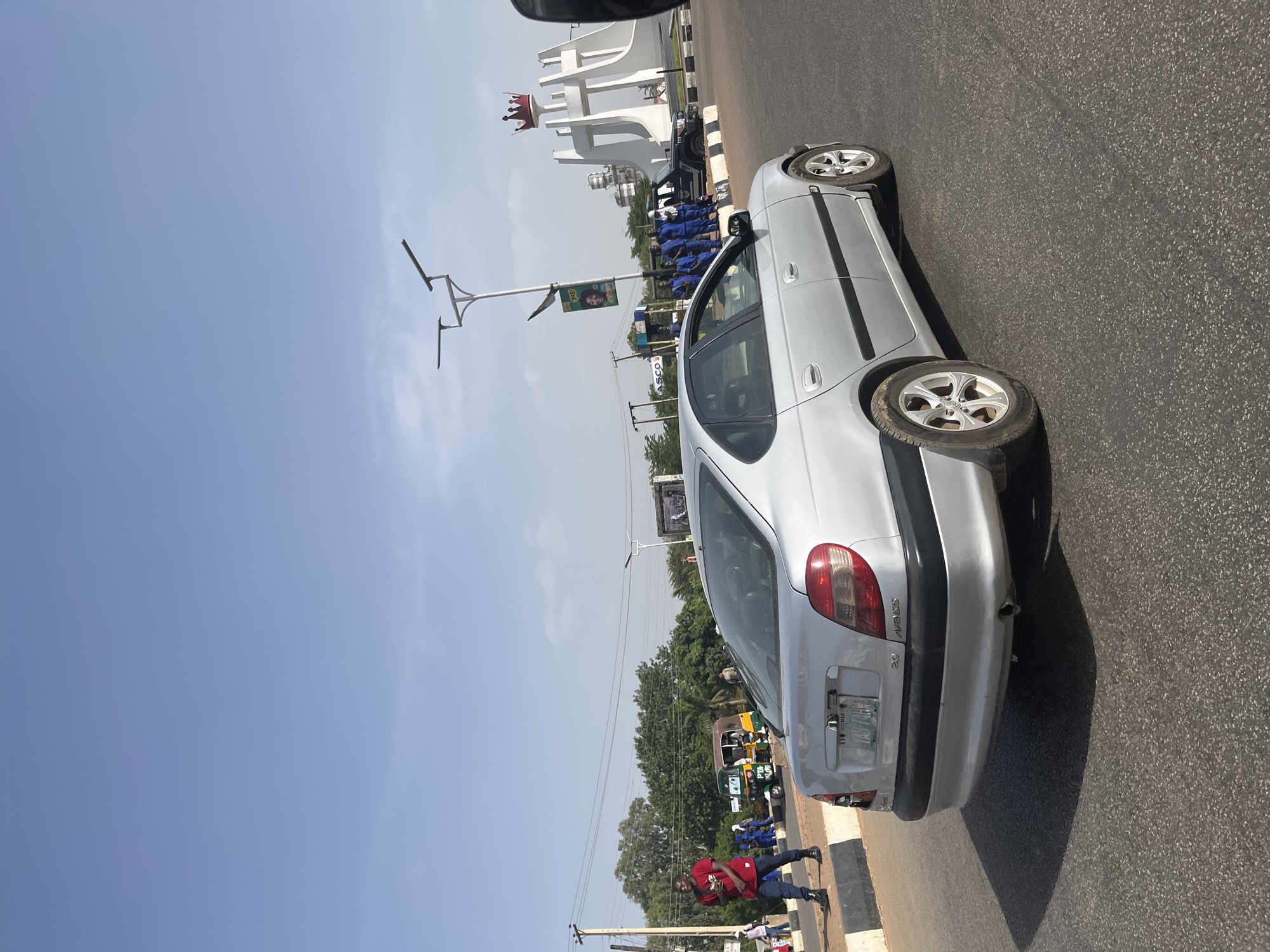 The image depicts a silver sedan, possibly a Peugeot 206, parked on the right side of a paved road in Jos, Nigeria. The road surface is asphalt. The rear of the vehicle is visible, showing the license plate. A group of uniformed individuals in blue are standing near a white sculpture with a large crown on top, which serves as a monument for Plateau State.
A man in a red shirt is walking on the road along a paved sidewalk, closer to the camera. Further down the road, there is a line of utility poles and green foliage. The sky is clear and blue, indicating a sunny day. There are other vehicles visible in the distance, including a yellow tricycle.