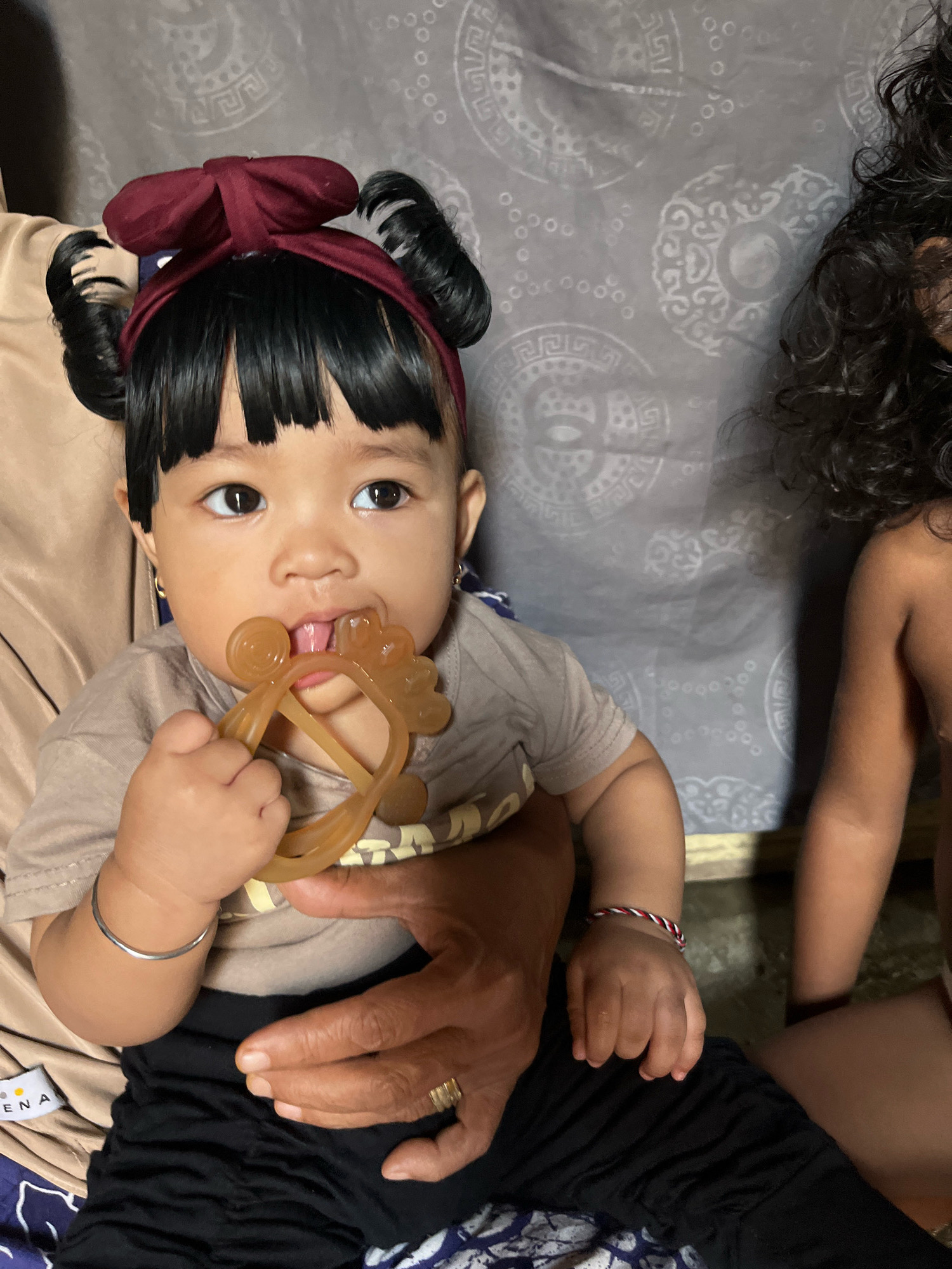 A close-up, eye-level shot captures a baby girl with dark hair styled in two buns and a maroon bow headband. She is wearing a light brown t-shirt and black pants. The baby is holding a light brown teething toy in her mouth and appears to be chewing on it. Her gaze is directed towards the viewer. The baby is seated on an adult's lap, with an adult's hands visible supporting her. To the right of the baby, partially visible, is another child with dark, curly hair. The background is a wall with a grey fabric covering featuring a repeating pattern of circular motifs and geometric shapes. The scene is dimly lit, creating a somewhat intimate and casual atmosphere. The image was taken in Kruengmate, Indonesia.