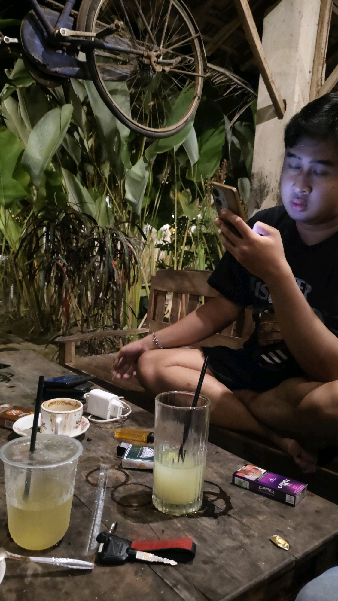 In Karangtalun, Indonesia, a young man is seated at a wooden table, engrossed in his smartphone. The table is cluttered with various items, including a plastic cup filled with a light-colored beverage and a straw, a ceramic cup with a saucer, a phone charger, a cigarette pack, a lighter, a glass of the same beverage, a cigarette, a car key, and a crumpled piece of foil. Behind the table, a wooden bench is visible. The background features lush greenery, with large leaves and some flowering plants. Suspended above the scene is an old bicycle wheel, adding an unusual element to the environment. The overall setting suggests a casual outdoor gathering, possibly at a cafe or a backyard in Karangtalun.