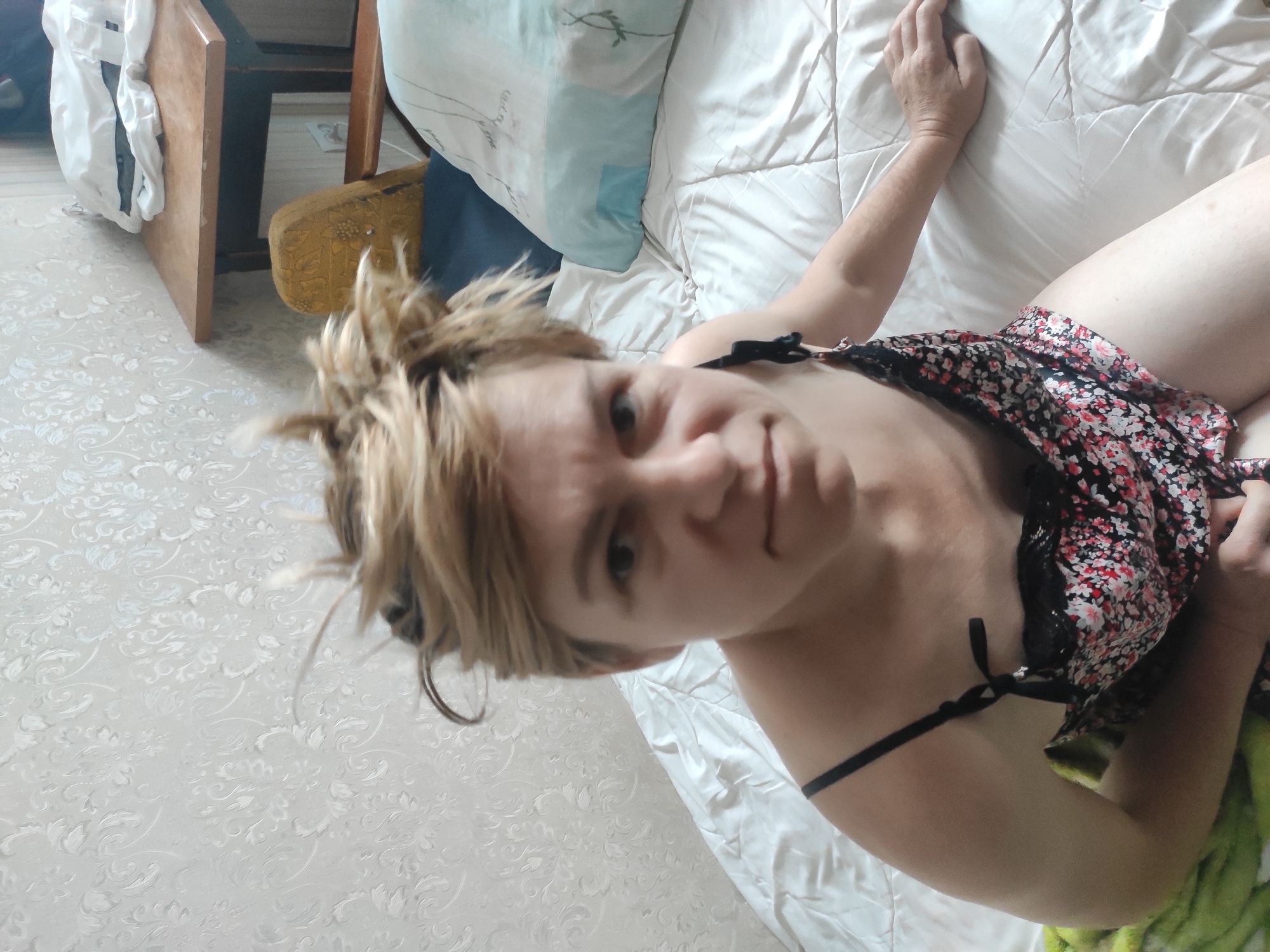 In a dimly lit bedroom in Kostopil, Ukraine, a person with short, spiky blonde hair is lying on a bed covered with a beige duvet. They are wearing a floral print slip dress with black straps. The person is looking directly at the camera with a serious expression. In the background, a bedside table holds a stack of white clothing, and a yellow upholstered chair is visible. The walls are covered with patterned wallpaper. A light green blanket is draped over the edge of the bed on the left side of the frame.
