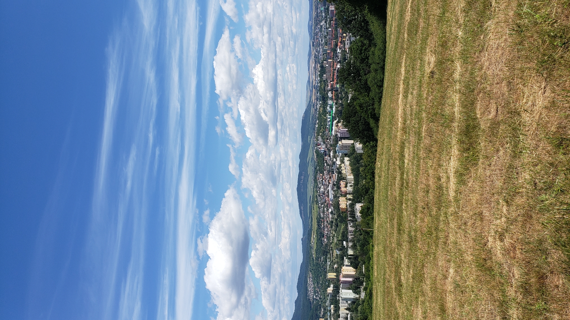 This is a vertical, eye-level shot of a panoramic view of the city of Prešov, Slovakia, taken from a grassy hilltop. The foreground is dominated by dry, straw-colored grass, indicating a summer or late summer season. In the mid-ground, a sprawling urban landscape unfolds, characterized by a mix of residential buildings, commercial structures, and scattered green spaces. The buildings are a variety of heights and architectural styles, with some appearing to be apartment blocks and others smaller, more varied structures. Trees and vegetation are interspersed throughout the city, softening the urban edges and creating a sense of integration with nature.
In the background, rolling hills and mountains form a natural backdrop to the city. The sky is a vivid blue, punctuated by scattered white cumulus clouds and thinner, wispy cirrus clouds that stretch across the horizon. The lighting suggests it is daytime, with the sun likely positioned to the right of the frame, casting subtle shadows and illuminating the scene. The overall impression is one of a peaceful, sunny day overlooking a populated area nestled within a picturesque natural setting. There are no people visible in the image.