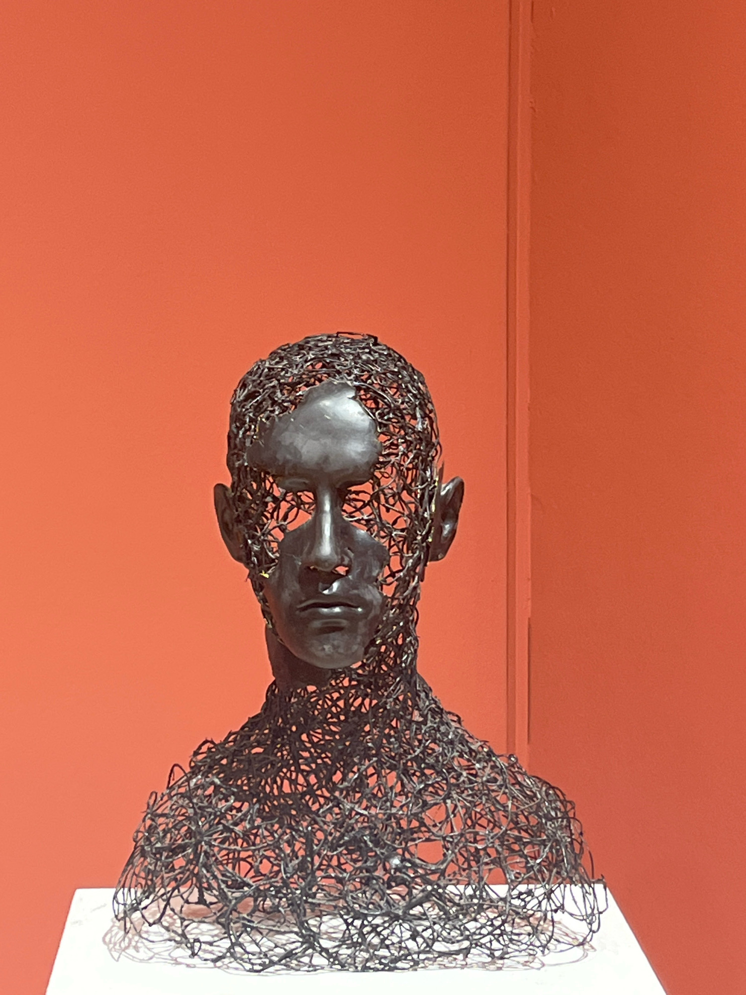 A wire sculpture of a human bust, made of dark, intertwined metal wires, is displayed against an orange wall. The sculpture has a solid, dark gray face with defined features, including eyes, a nose, and lips. The wire mesh forms the head and neck, with gaps revealing the space behind the sculpture. The bust is placed on a white pedestal. The background is a plain, solid orange wall with a vertical seam visible on the right side, suggesting a corner. No people are present in the frame. This artwork is located in Podgorica, Montenegro.