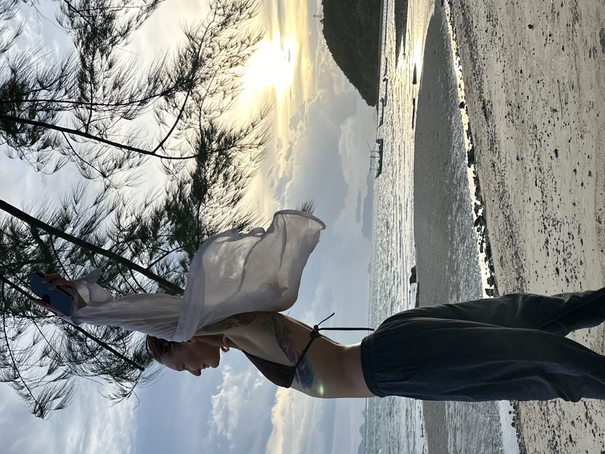 The image, captured in the Ko Samui District of Thailand, depicts a person standing on a sandy beach near the water's edge. The sun is setting, casting a warm golden glow across the sky and reflecting on the calm sea. The sky is filled with scattered clouds, illuminated by the low-lying sun. Silhouetted against the dramatic sky are trees with wispy needles, their branches reaching out towards the viewer. In the background, a lush green island rises from the sea. Small boats are visible in the water, hinting at local fishing or transport activities.
The person, viewed from the side, is wearing dark pants and a bikini top. A sheer white garment, possibly a shirt or scarf, is held aloft by their arms, billowing in the gentle breeze. The fabric is captured mid-air, creating a dynamic and ethereal effect. Tattoos are visible on the person's arms and torso. They are holding a blue smartphone in their left hand, seemingly taking a picture or video. The overall mood is serene and contemplative, with a sense of freedom and connection to nature. The bright, diffused light suggests the golden hour just before sunset.
