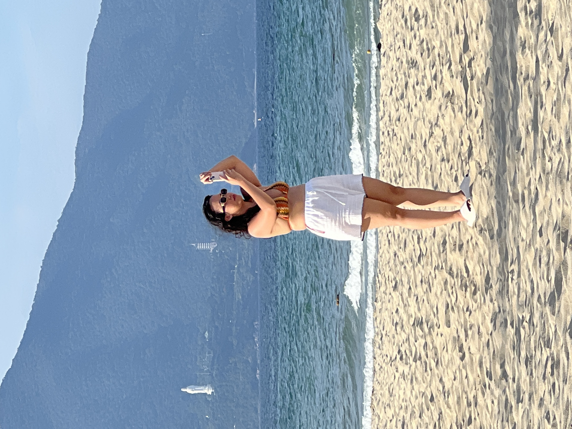 A woman stands on a sandy beach, holding a smartphone to take a picture or video. She is wearing a striped bikini top and white shorts, along with sunglasses and white slides. Behind her, the ocean stretches out, with gentle waves lapping the shore. In the distance, a large, green mountain dominates the horizon, with a white statue of a goddess and a tall, modern-looking building visible on its lower slopes. The beach itself is composed of light-colored sand with visible footprints and shadows. The sky above is clear and blue. This scene is likely the beach at Sơn Trà Peninsula in Đà Nẵng, Vietnam, given the presence of the Hải Vân Pass mountains and the Goddess of Mercy statue.