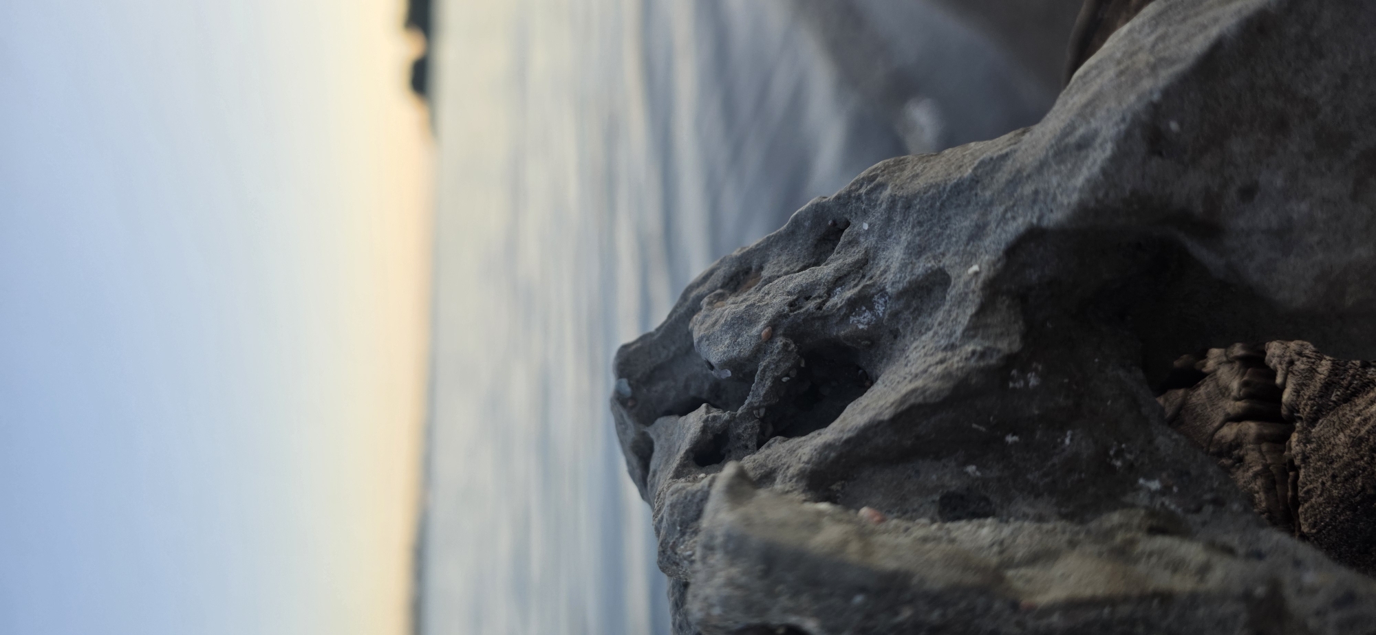 The image depicts a close-up of a weathered, porous rock formation in the foreground. The rock is gray and textured, with several natural openings and irregularities. Behind the rock, a body of water stretches to the horizon, reflecting the soft colors of a twilight sky. The water appears calm with subtle ripples. In the distance, a blurry landmass, possibly a shoreline with faint silhouettes of structures or trees, is visible against the horizon. The sky transitions from a pale blue at the top to a warm, soft yellow-orange glow near the horizon, indicating either sunrise or sunset. There are no people, animals, or any discernible activity in the image. The scene appears to be a natural landscape. The location is stated to be Issyk-Kul, Kyrgyzstan.