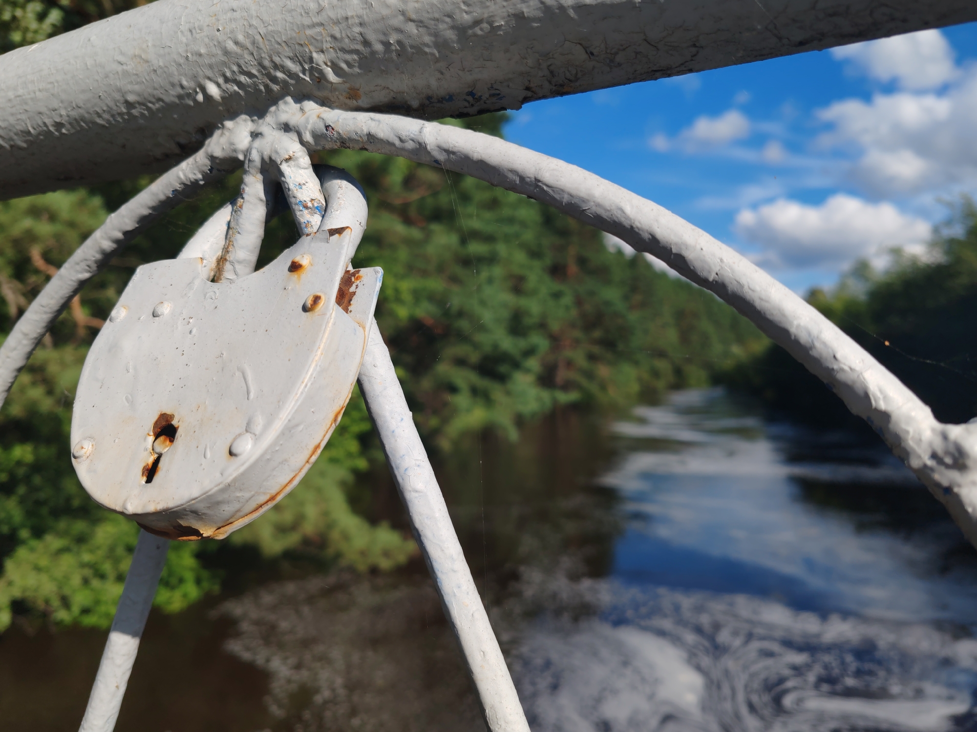 The image shows a close-up of a weathered, light gray padlock hanging from a white, painted metal railing. The padlock, which appears old and rusty in places, is attached to two curved metal bars that are part of the railing. In the background, a body of water, possibly a river, flows through a densely wooded area. The water has some swirling patterns on its surface. The sky above is blue with scattered white clouds. The overall scene suggests a bridge or overlook in a natural setting, likely in the vicinity of Кашино, Russia, given the context. No people or specific actions are depicted.