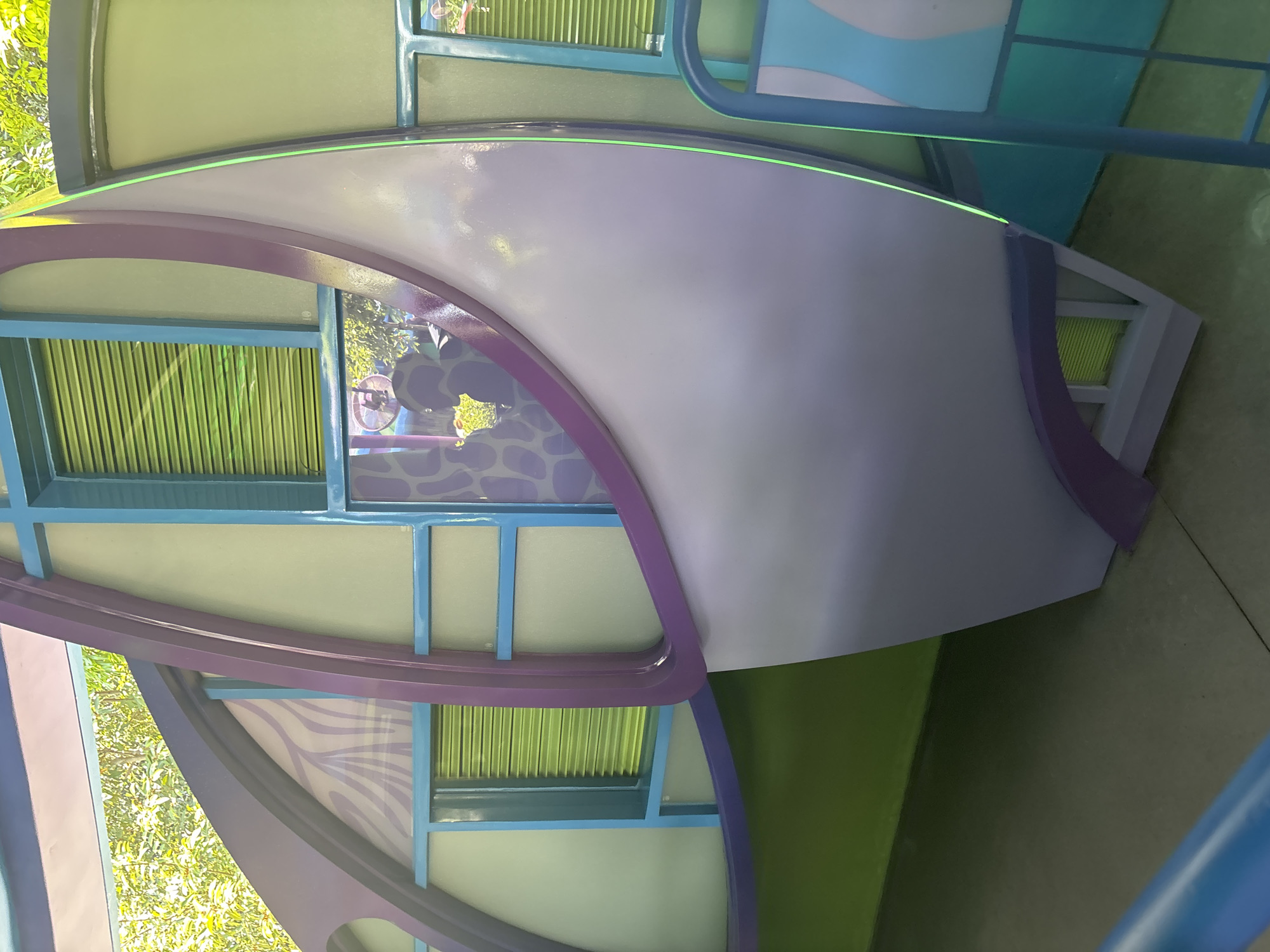 The image shows a colorful, whimsical structure with a curved, light purple panel dominating the foreground. The structure features blue framing and translucent, light green panels behind slatted green windows. A large purple arch frames a section of the structure. In the background, green foliage is visible, suggesting an outdoor setting. A portion of a blue railing is visible in the lower right corner. The scene appears to be located in Pudong, China, possibly in an amusement park or public art installation, given its playful design. There are no people or specific actions depicted.