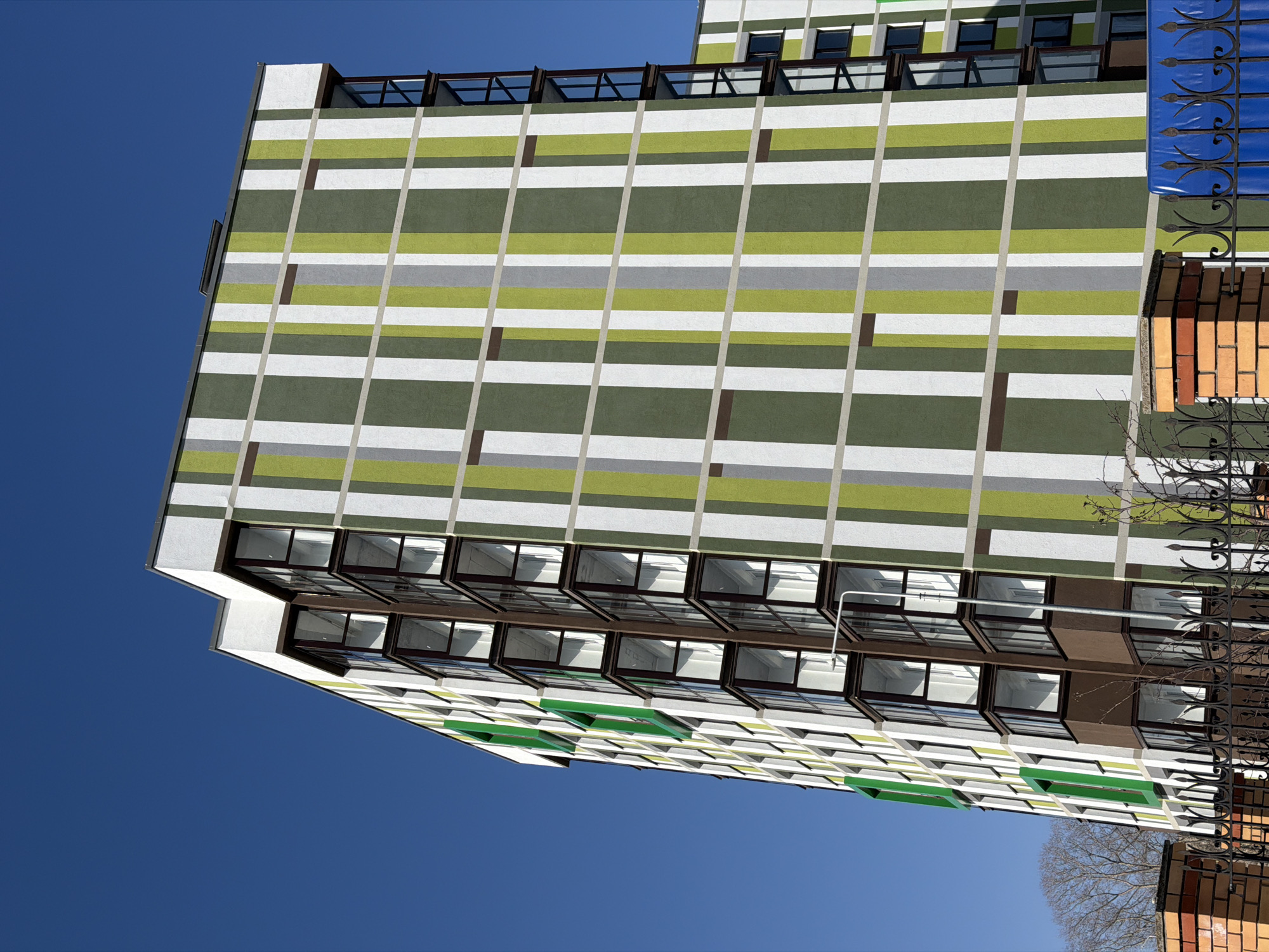 An upward-angled shot reveals a modern apartment building under a clear, blue sky in Berezniki, Russia. The building's facade features a distinctive, patterned design of horizontal stripes in alternating shades of green, white, and gray, divided by vertical lines. Rectangular, dark brown accents punctuate the striped pattern. Below, numerous windows are visible with dark frames, some partially open. A wrought-iron fence and a brick structure appear in the lower right corner of the frame, suggesting the building's proximity to other structures. A few bare tree branches are also visible.