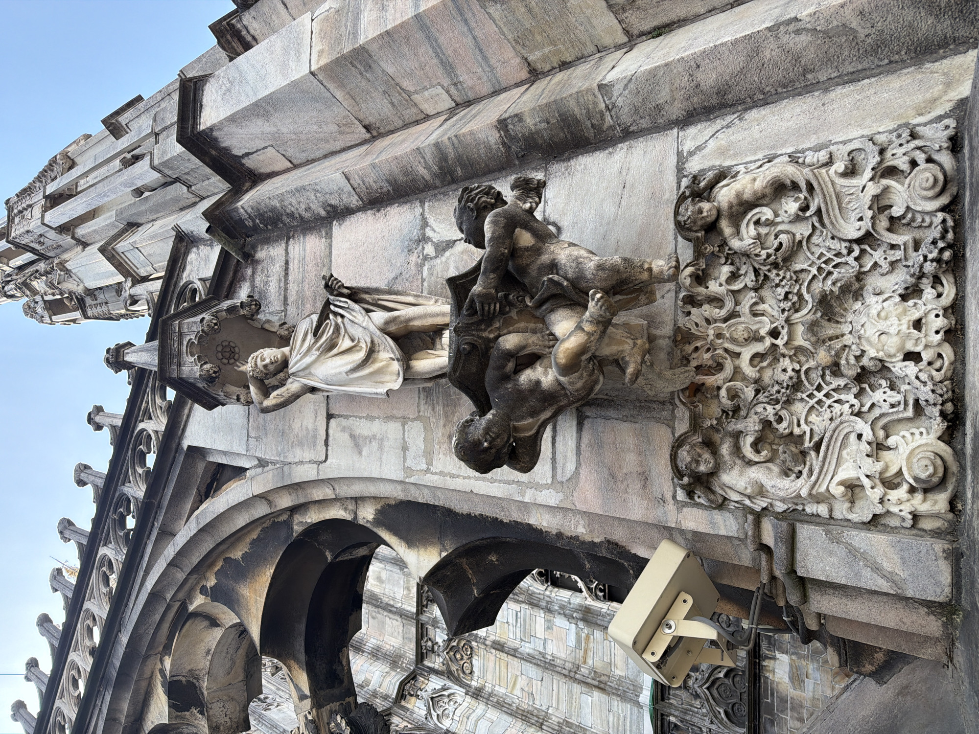 This upward-angled, outdoor shot depicts a section of the Duomo di Milano in Milan, Italy. The image highlights architectural details, including stone carvings and structural elements.
On the right side of the frame, a rectangular carving featuring intricate floral and scroll patterns is visible. Adjacent to this, a sculpture depicting two figures, one of whom has wings, rests atop a larger architectural element. To the left, there is an arched window or doorway section with dark, weathered stone. Above the archway, a series of vertically oriented stone structures with decorative pinnacles ascend towards the sky.
The composition is largely static, emphasizing the texture and detail of the building's stonework. In the foreground, a security light fixture is attached to the building. The sky is visible in the upper-left corner, appearing bright and clear.