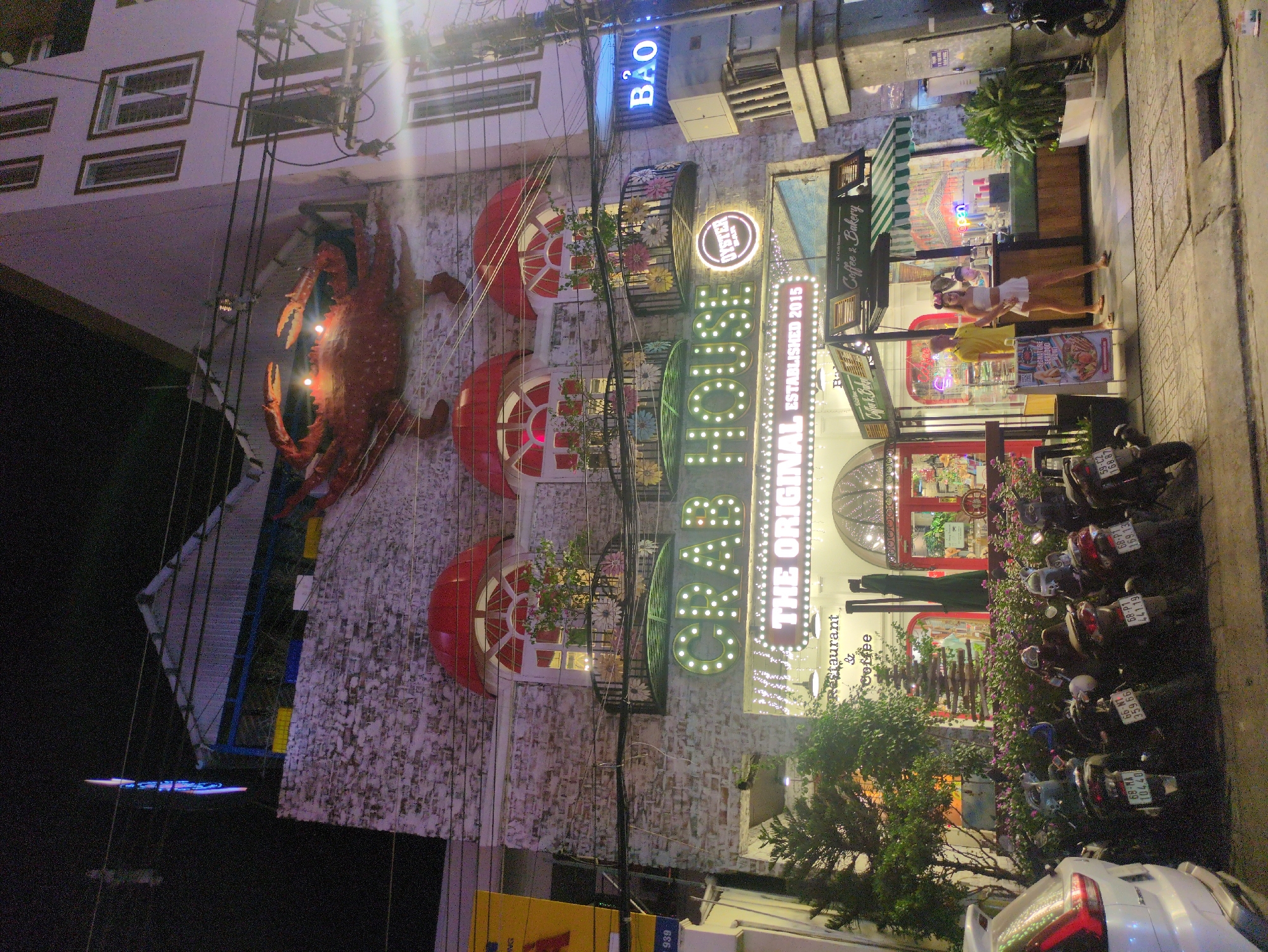 The image captures a nighttime street scene in Phú Quốc, Vietnam. The focal point is a restaurant named "Crab Bar," which features a large crab decoration on its facade. The building's exterior has three red-trimmed windows, and signs advertising "The Original Established 2015," "Oyster Bar," and "Coffee & Bakery." Two individuals are standing in front of the establishment, with one wearing a yellow shirt and the other a white top and shorts. Parked motorbikes line the curb in front of the restaurant. The street appears well-lit. A sign with the word "BẢO" is visible on the building.