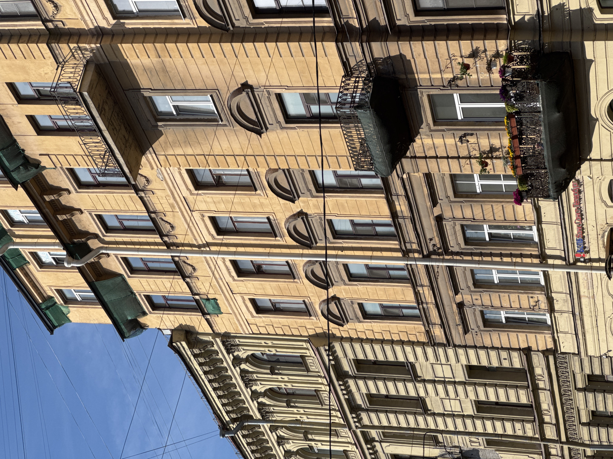 This is a medium-angle, eye-level, exterior, daytime, vertical, full-shot photograph of the facades of two adjacent, multi-story buildings in Saint Petersburg, Russia. The buildings are made of a textured, ochre-colored stucco. The building on the right has a balcony with an ornate black metal railing and a green tarp underneath, containing flower boxes with colorful flowers. The building on the left has a more elaborate facade with arched windows and decorative columns. Electrical wires crisscross the scene against a clear blue sky. The buildings appear to be old, with some signs of wear on the stucco. There are no people visible in the image. The architectural style suggests a historical district in Saint Petersburg.