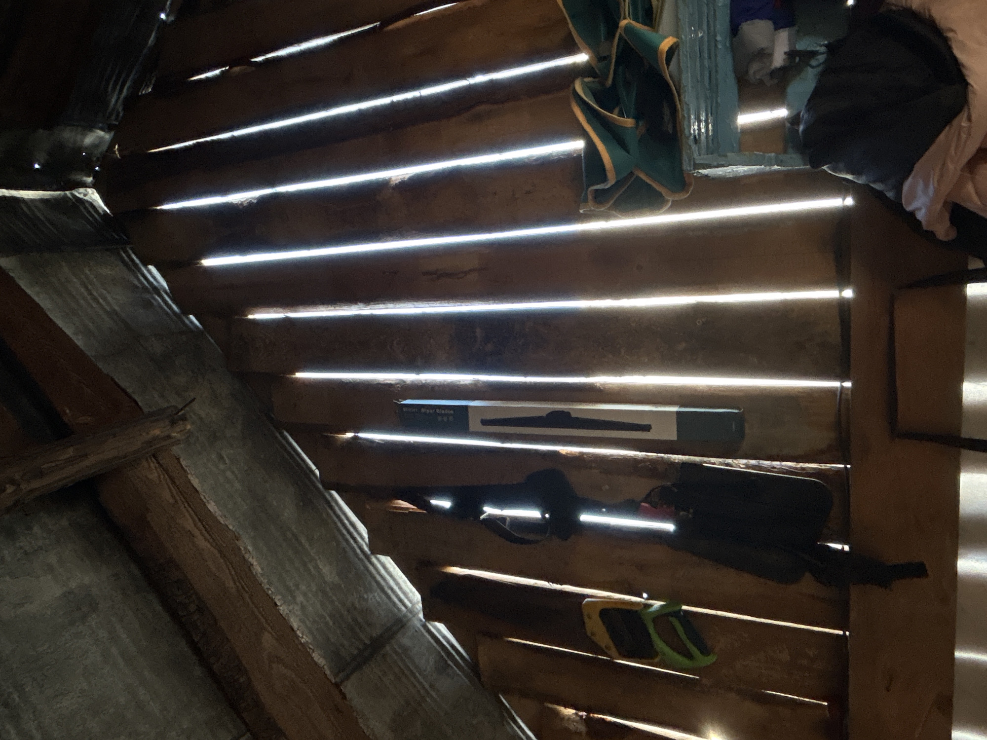 The image shows the interior of a wooden structure in Chaikovsky, Russia. Light streams through gaps in the wooden boards that form the walls. Several objects are visible, including a box labeled "Super Wiper Blades", a dark-colored object that may be a tool, and green flip-flops. Clothes hang from the structure above. The wood appears aged and weathered.