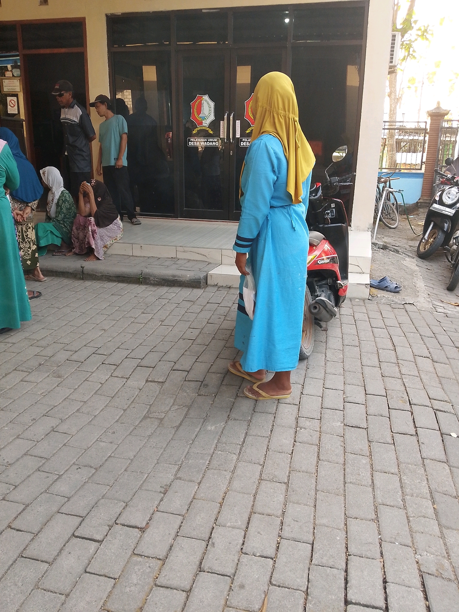 In front of the village hall of Wadang, Indonesia, a group of people are gathered. A woman in a blue robe and yellow headscarf stands facing away from the camera, holding a plastic bag. Several other people are seated on the steps of the building, including a woman in a green and white patterned dress and a woman in a dark hijab and patterned skirt, who is covering her face. Two men stand near the entrance, one in a dark shirt and cap, and the other in a light blue t-shirt and a baseball cap. Motorcycles are parked on the paved area in front of the building. The ground is covered in grey brick pavers, and a potted plant is visible to the right of the entrance. The building has large glass doors and a sign that reads "PELAYANAN UMUM DESA WADANG". The scene appears to be a public service area within the village.
