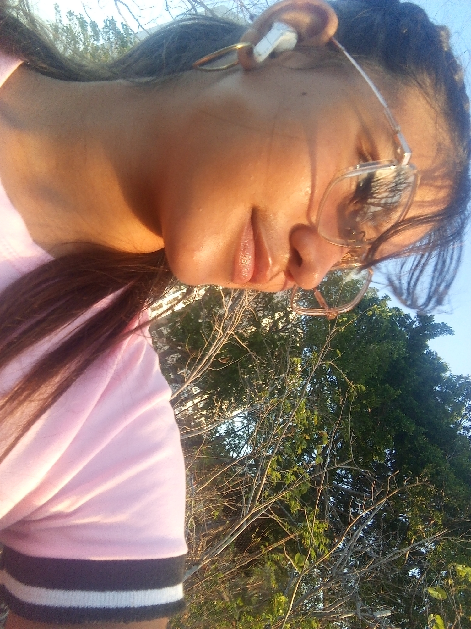 A low-angle selfie shows a female individual wearing clear-rimmed glasses and a white wireless earbud in their right ear. The person has brown hair and is wearing a pink shirt with black and white stripes on the sleeve. The background reveals trees and a blue sky, suggesting an outdoor environment. It is impossible to determine whether this photo was taken in the city of Juan Griego, Venezuela.
