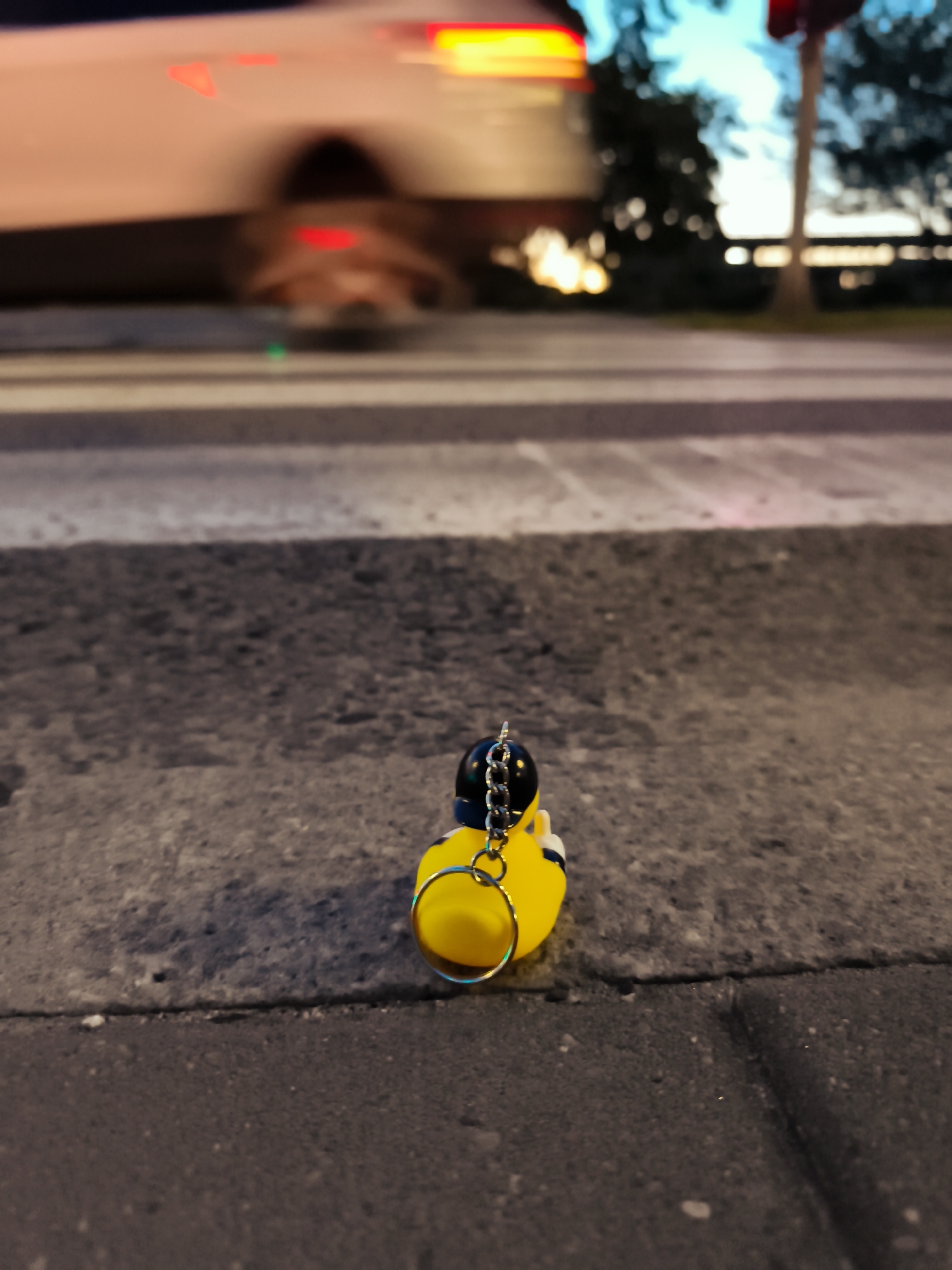 A close-up, low-angle shot depicts a yellow rubber duck keychain resting on a gray asphalt surface, likely a crosswalk, in Gdańsk, Poland. The duck, adorned with a black hat and silver chain, faces to the right. In the blurred background, a white vehicle with illuminated taillights is in motion, suggesting a street scene at dusk or night. Trees and a bright sky are also visible, contributing to the urban setting.