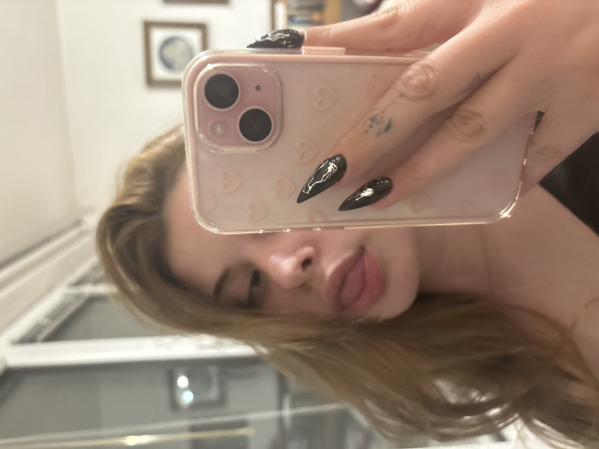 The image is a selfie taken with an iPhone encased in a pink phone case adorned with a pattern of hearts. The phone obscures a portion of the subject's face, focusing on the mouth with the tongue slightly protruding. The subject has light brown hair and black nails with white designs. The background appears to be an interior space, possibly a house, with windows and framed artwork visible. The location is not definitively discernible as Troyan, Bulgaria. The information is not provided in the image.