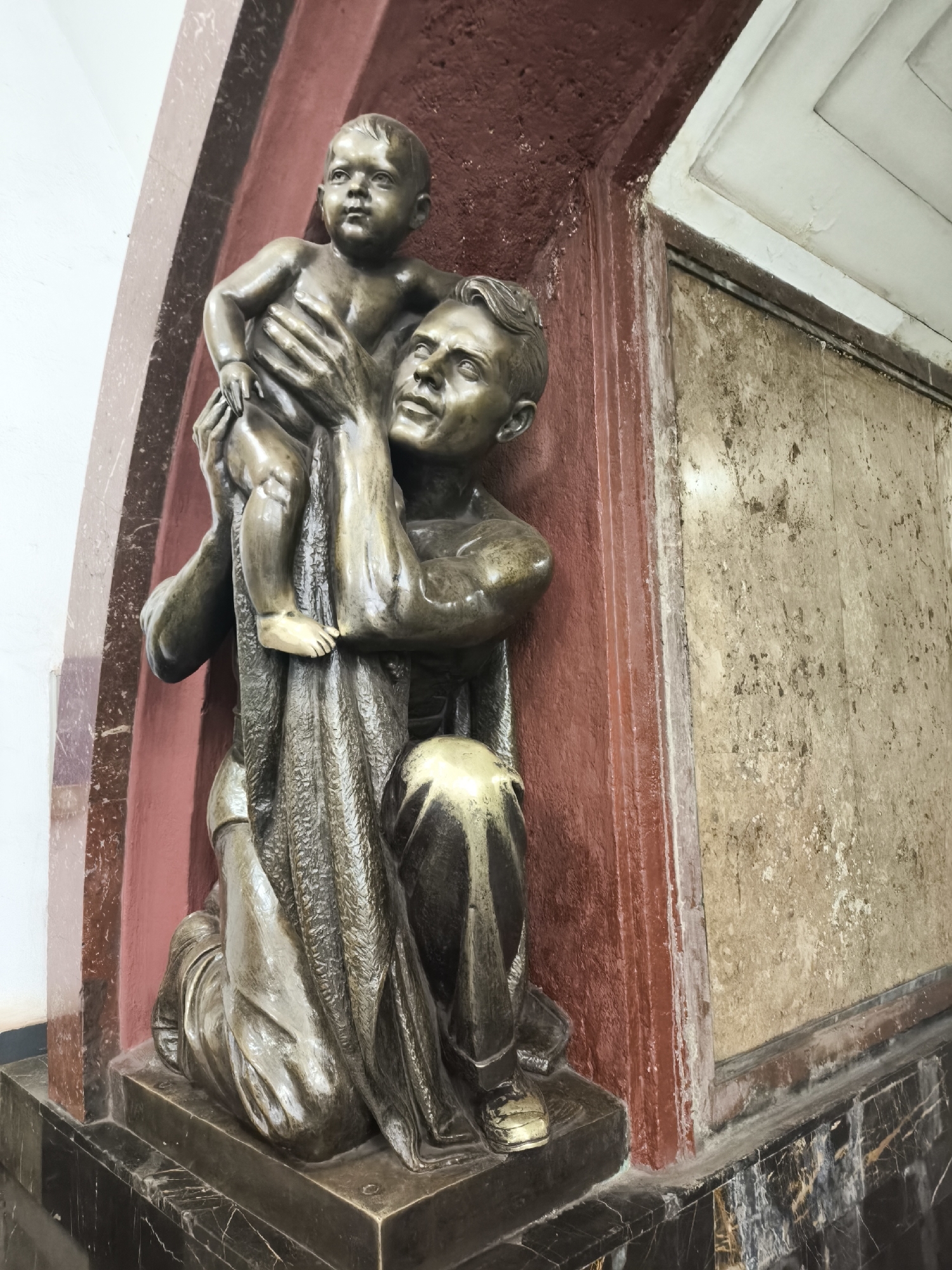 A statue of a man holding a child is affixed to a wall. The statue is made of bronze and the man is kneeling, holding the child up in his arms. The child is looking forward with a serious expression. The man is looking up at the child with a smile. The wall is red and white, and the statue is standing on a black marble base. The location is in the city of Unknown, Unknown.