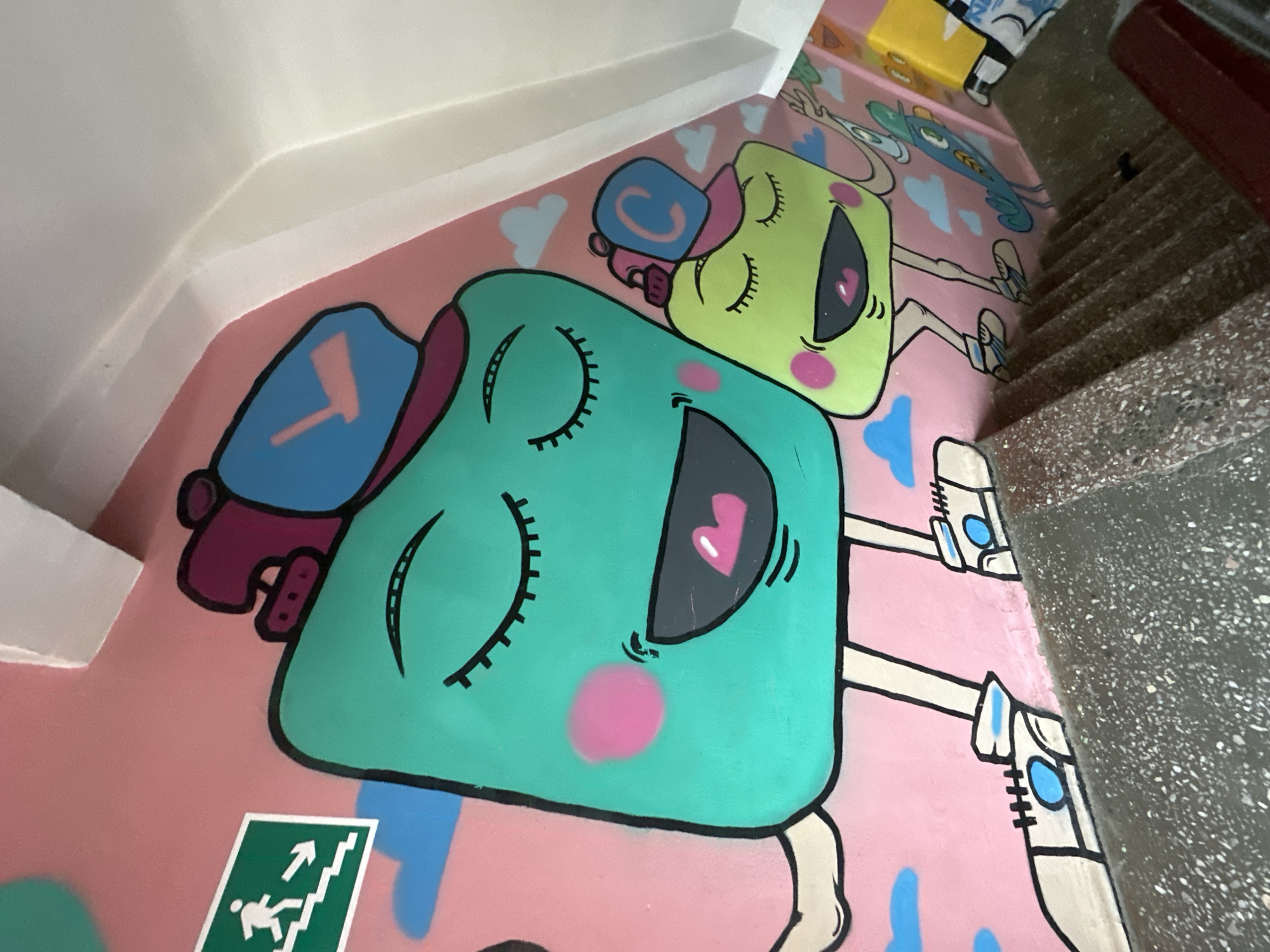 A mural is painted on a pink wall inside a building, likely a stairwell, in Biysk, Russia. The mural depicts two cartoon characters, one larger teal-colored figure with closed eyes, rosy cheeks, and an open mouth, wearing a purple and blue hat. Beside it is a smaller, lime green figure with similar features and a pink and blue hat. Both characters have thin arms and legs and are wearing white sneakers with blue accents. The wall also features painted blue clouds. A green emergency exit sign with a running person and arrows is visible to the left of the larger figure. The image is taken from a high angle, looking down a set of concrete stairs.