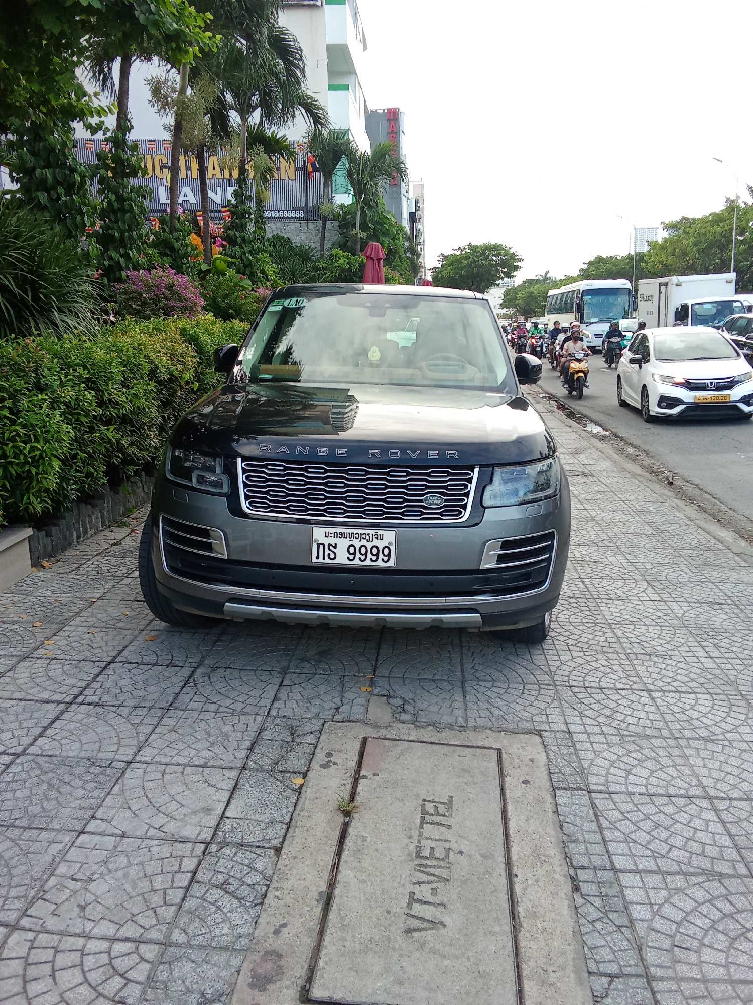 The image shows a black Range Rover parked on a sidewalk in Da Nang, Vietnam. The car is facing the camera, and its license plate reads "NS 9999". In the background, there is a busy street with traffic, including motorbikes, cars, and a bus. There are also trees and buildings lining the street. The sidewalk is paved with gray tiles. A metal manhole cover with "VT-VIETTEL" written on it is visible in the foreground. The overall scene depicts a typical urban street in Vietnam.