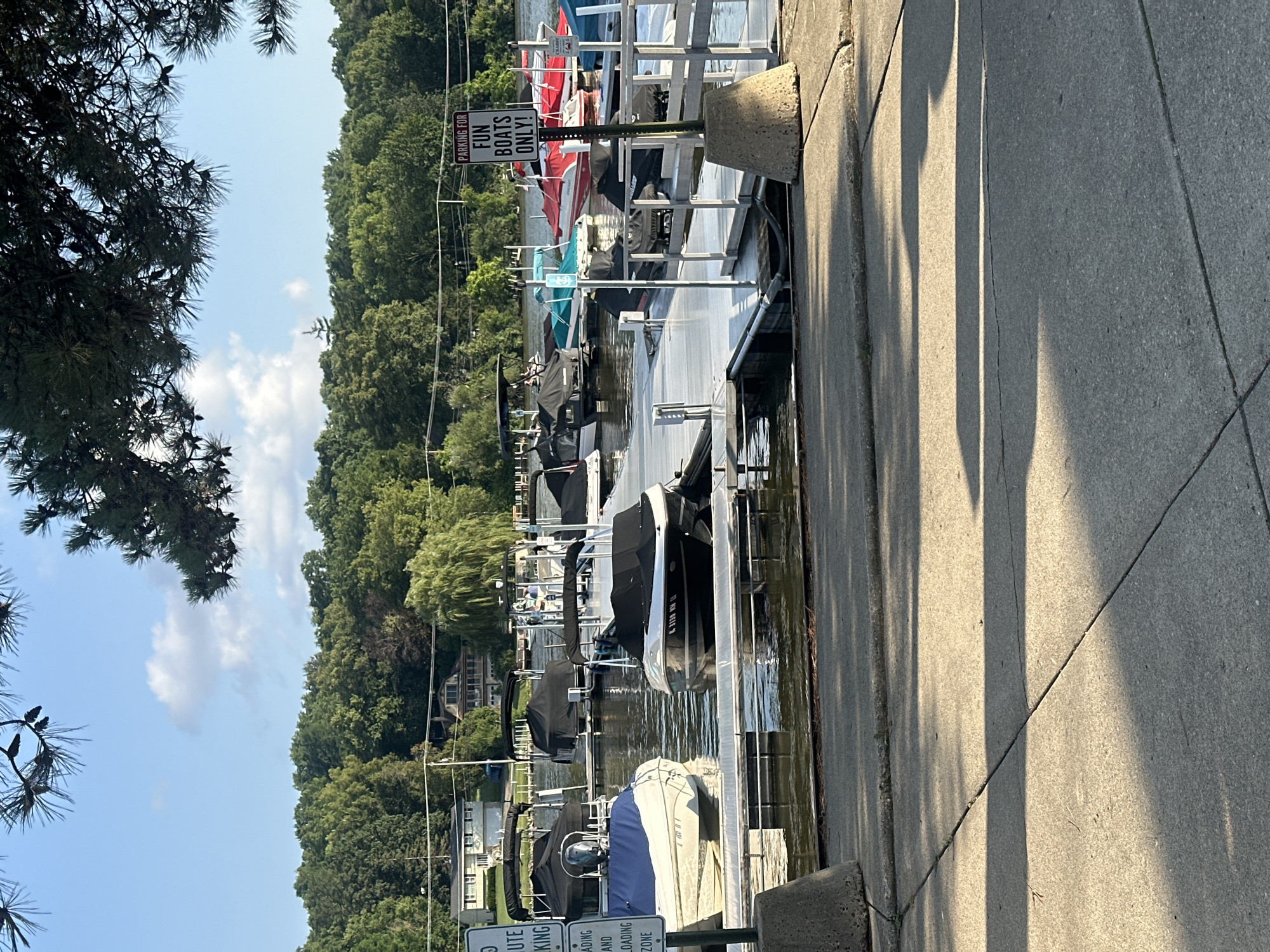The image captures a scene at a marina on the Fox River in Algonquin, Illinois, United States of America. The foreground shows a concrete walkway with shadows from overhead foliage. Beyond the walkway is a dock with several boats moored. Some boats are covered with tarps, while others are open. The boats vary in size and type, including what appear to be jet skis and smaller motorboats. A sign with the text "PARKING FOR FUN BOATS ONLY!" is visible on a post. In the background, a lush green treeline borders the river, with houses and buildings peeking through the trees. The sky is blue with scattered white clouds. The water of the river is calm, reflecting the surrounding greenery and sky. There are no visible people actively engaged in any activity in the image.