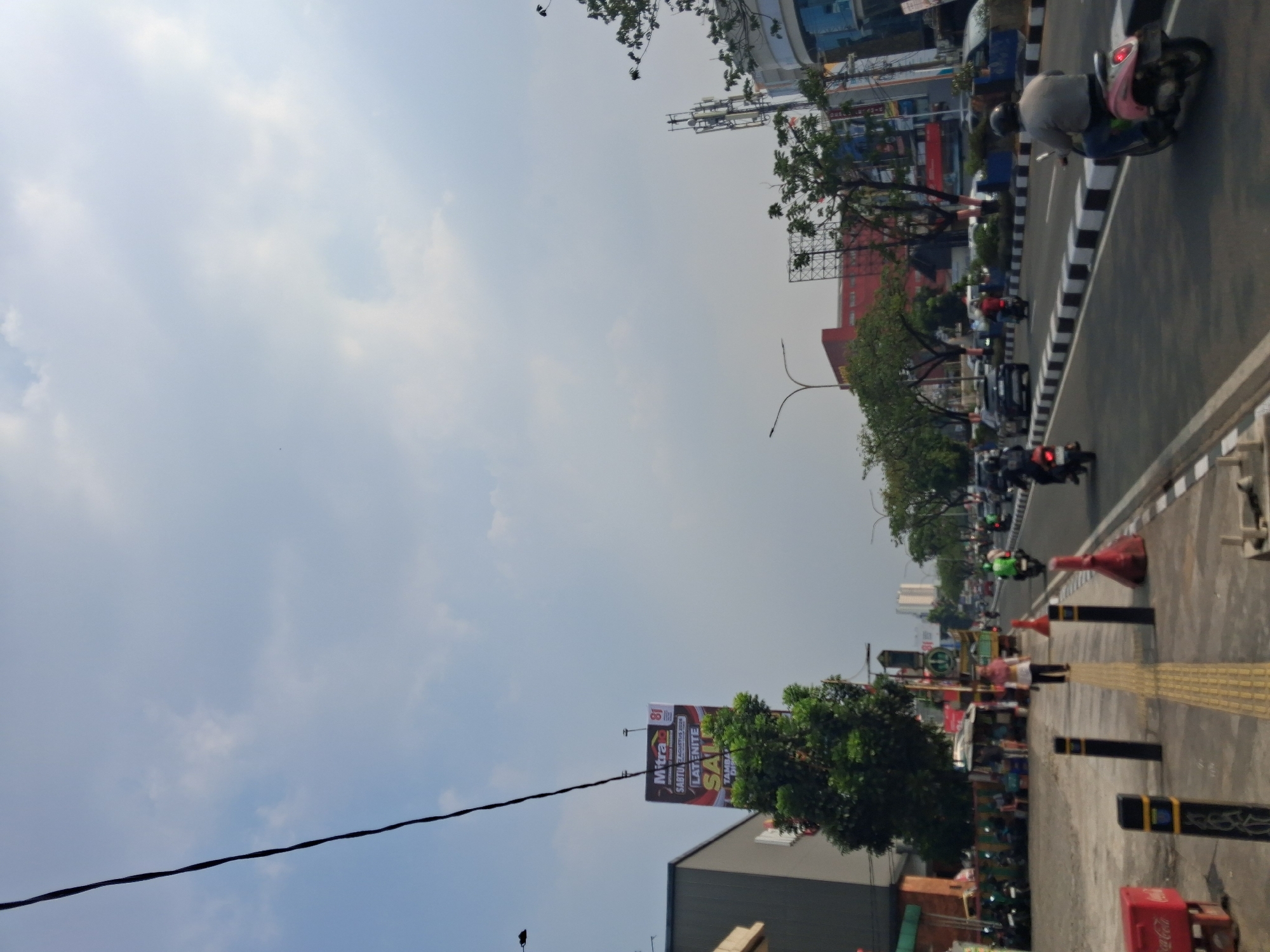 The image depicts a street scene in Depok, Indonesia, under a partly cloudy sky. A prominent billboard on the left advertises "Mitra 81" and "LATENITE SALE," with "SABTU 2 AGUSTUS 2020" visible. A dense green tree grows beside the billboard. The street is divided by a white and black striped curb. Multiple vehicles, including cars and motorcycles, are present on the road. Several people are visible, some walking on the sidewalk, others riding motorcycles. On the right side of the street, there are buildings with modern architecture and telecommunication towers. A pink scooter with a rider in a grey shirt is moving away from the camera on the right lane of the road. A red Coca-Cola cooler is on the sidewalk to the left. The overall atmosphere suggests a typical day in an urban environment with commercial activity.