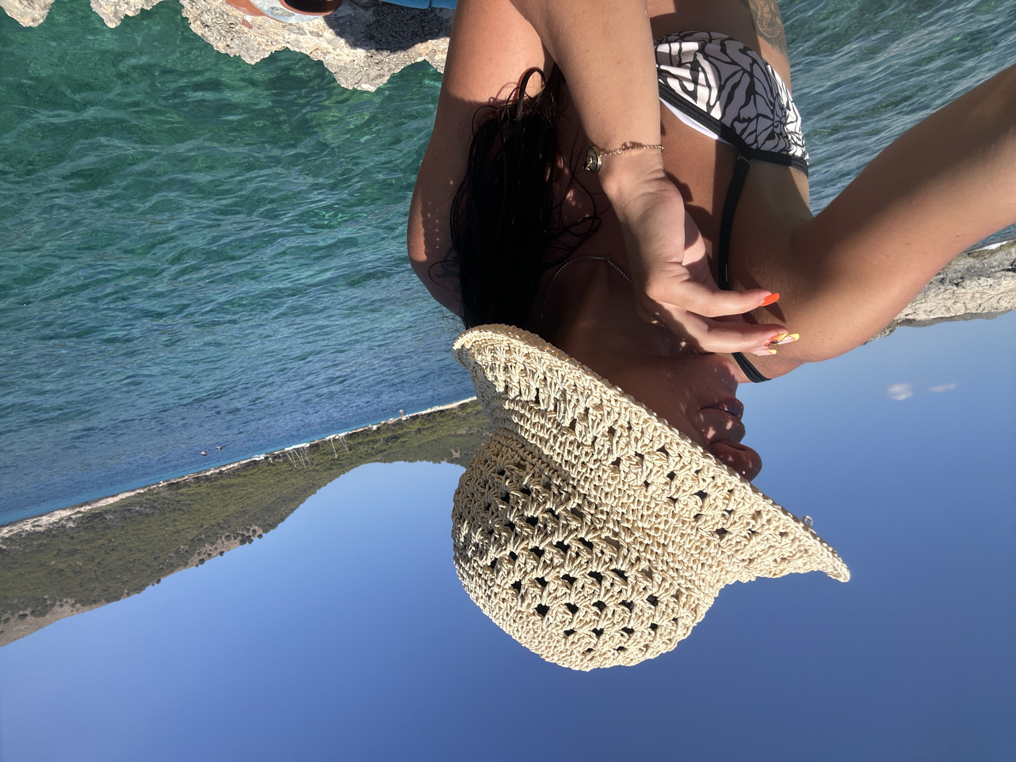 A person wearing a straw hat and a bikini poses for a selfie by the sea. The person is sitting on rocks overlooking clear turquoise water and a lush green hillside. The sun is shining brightly, casting shadows on the person and the rocks. The person's hand is resting on their chin, and their fingernails are painted with colorful designs. Sailboats are visible in the distance. The location is Cala Lliteres, Spain.