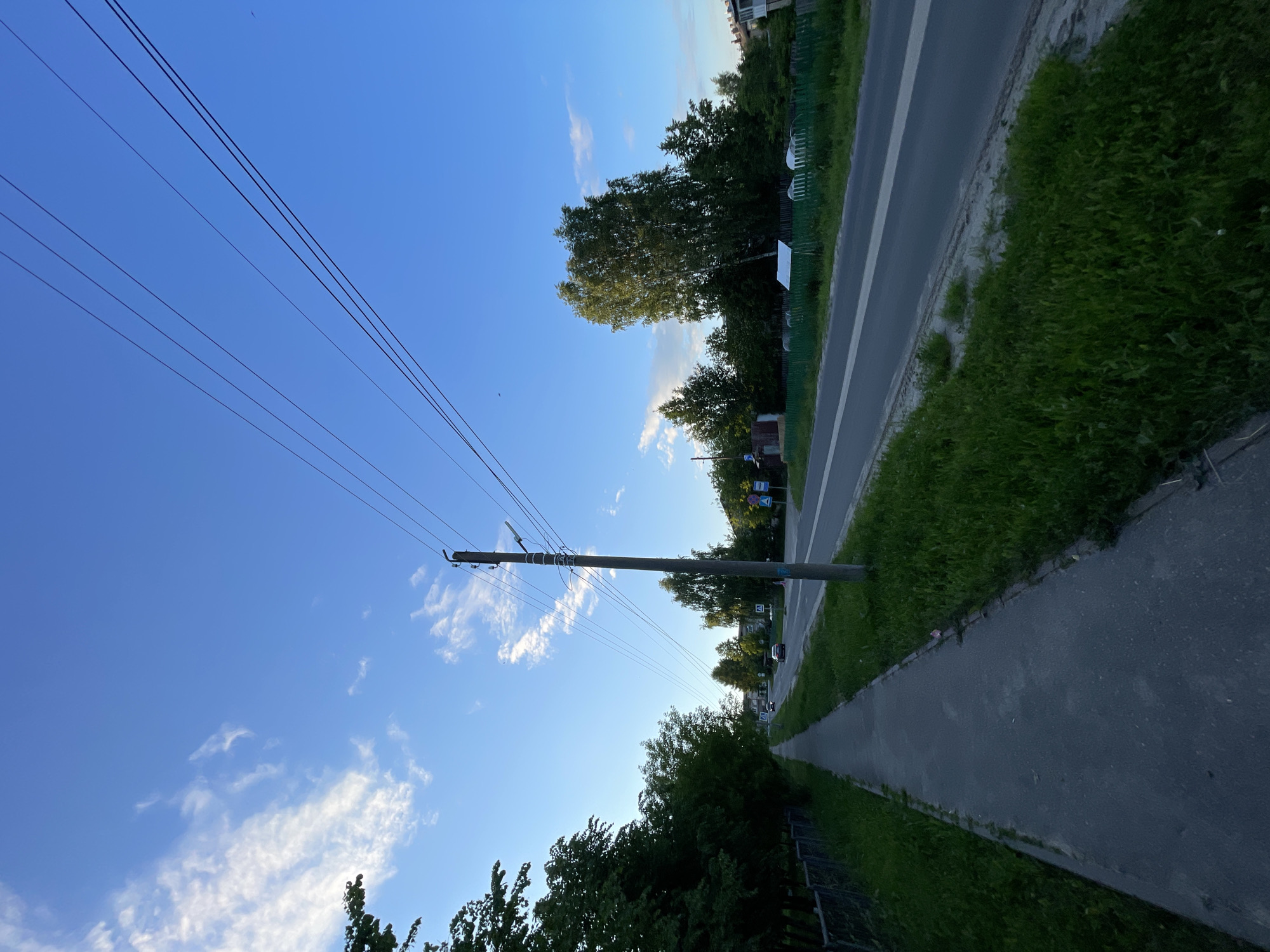 The image depicts a suburban street scene in Krasnye Baki, Russia, under a clear blue sky with scattered white clouds. A paved sidewalk runs along the left side of the frame, bordered by a grassy verge and a metal fence. On the right, a two-lane asphalt road is visible, with a white line marking the center. Several cars are present on the road, traveling in both directions. Utility poles with numerous wires stretch across the upper portion of the image. Lush green trees line both sides of the road, with some houses and fences visible in the background. The overall impression is of a quiet, residential area during daylight hours.