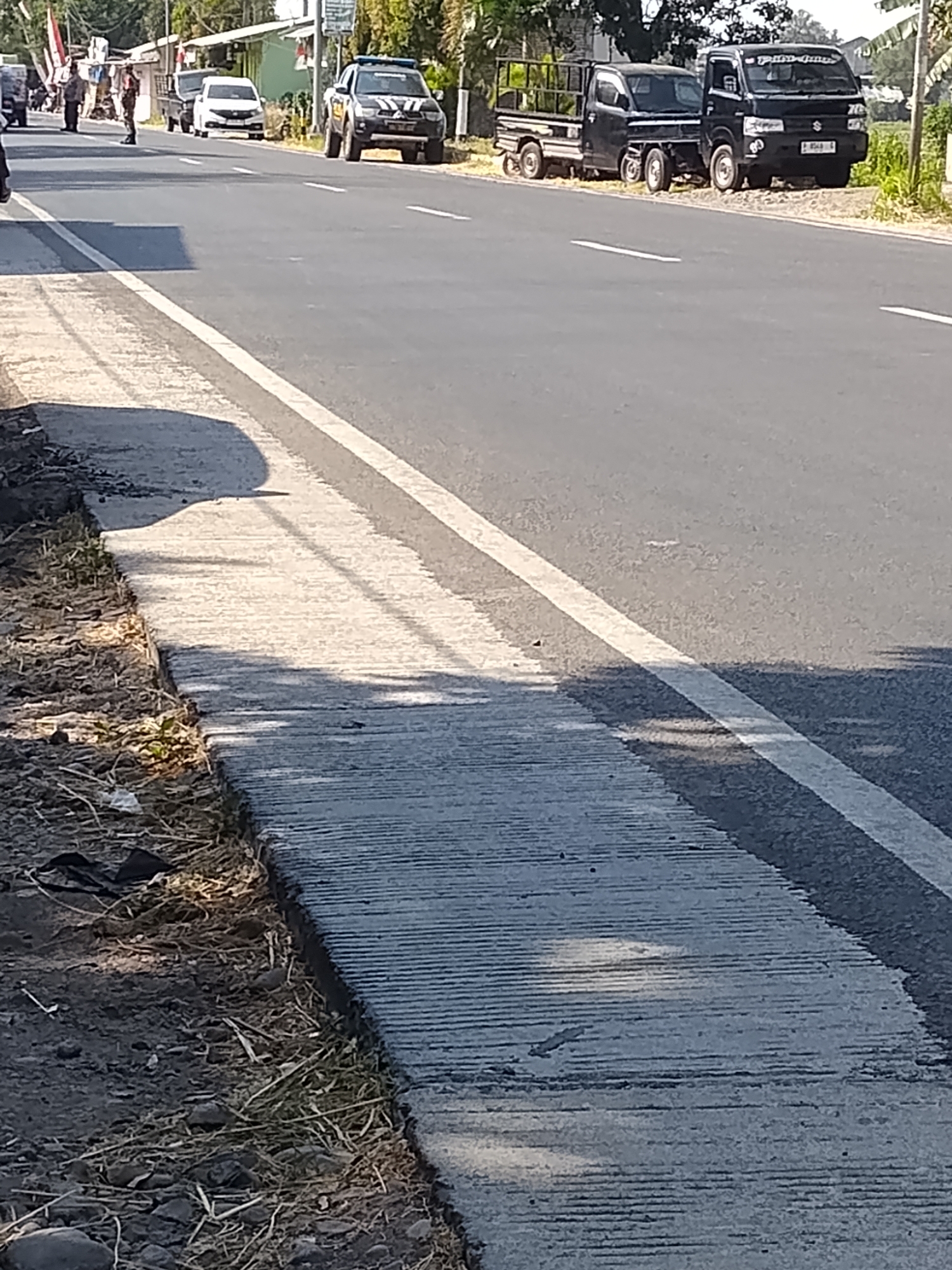 This is a street-level, medium-shot photograph taken in Buduan, Indonesia. The foreground shows a concrete sidewalk with a rough texture, bordered by dirt and debris. A white line marks the edge of the asphalt road, which occupies the midground. Several vehicles are visible on the road. On the far right, a black pickup truck is parked, followed by another black pickup truck. Further down the road, a white sedan is parked, and a black pickup truck with a cage in the back is also present. Two individuals, one in a dark uniform and another in a lighter-colored uniform, stand on the sidewalk near the white sedan. The background features buildings with awnings and trees. The lighting suggests it is daytime.
