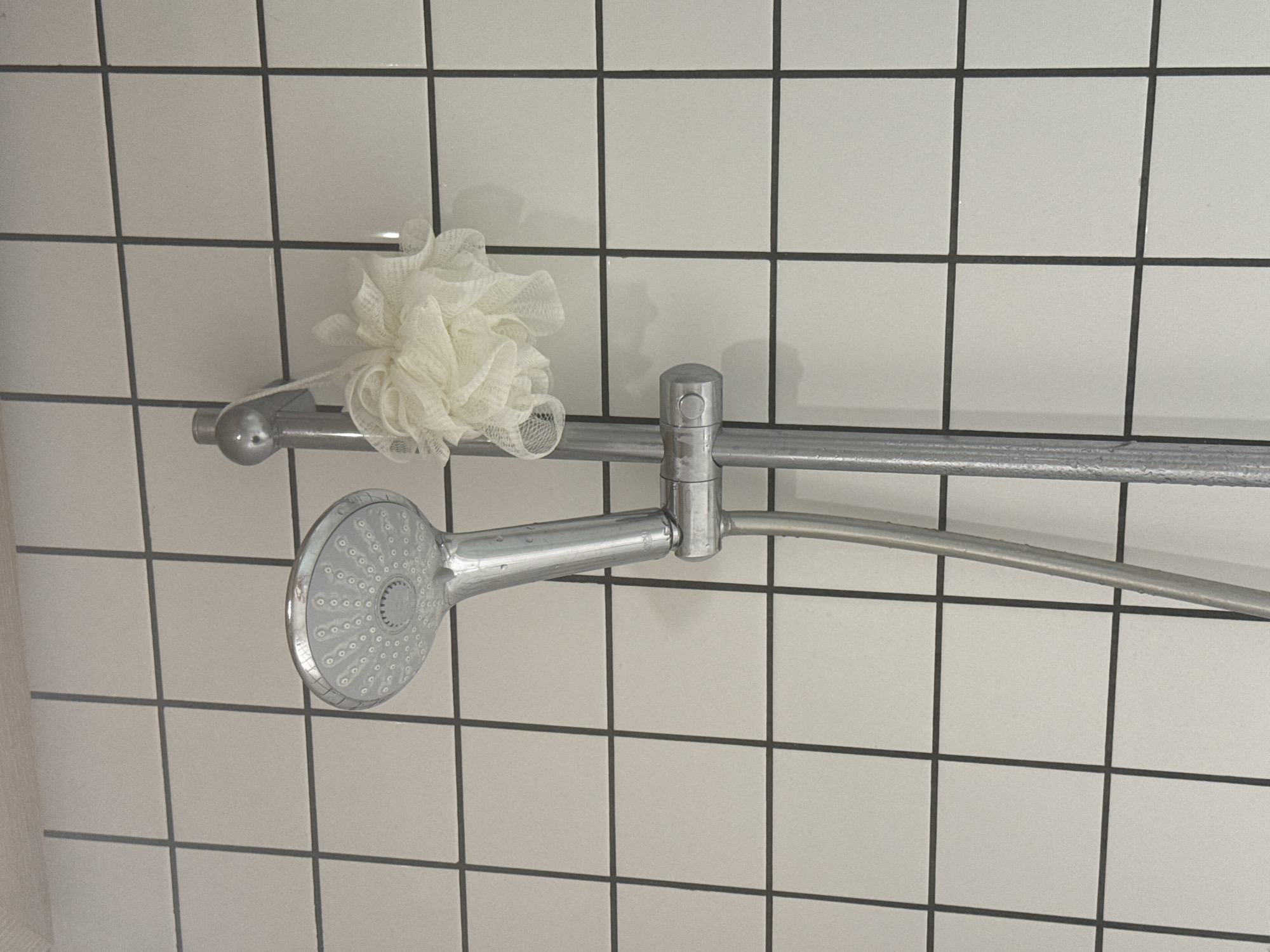 A chrome shower head is mounted on a chrome bar attached to a white tiled wall with black grout lines. A white mesh shower pouf hangs on the shower bar to the left of the shower head. The scene appears to be in a bathroom. The location is unknown and cannot be determined based solely on the provided image.