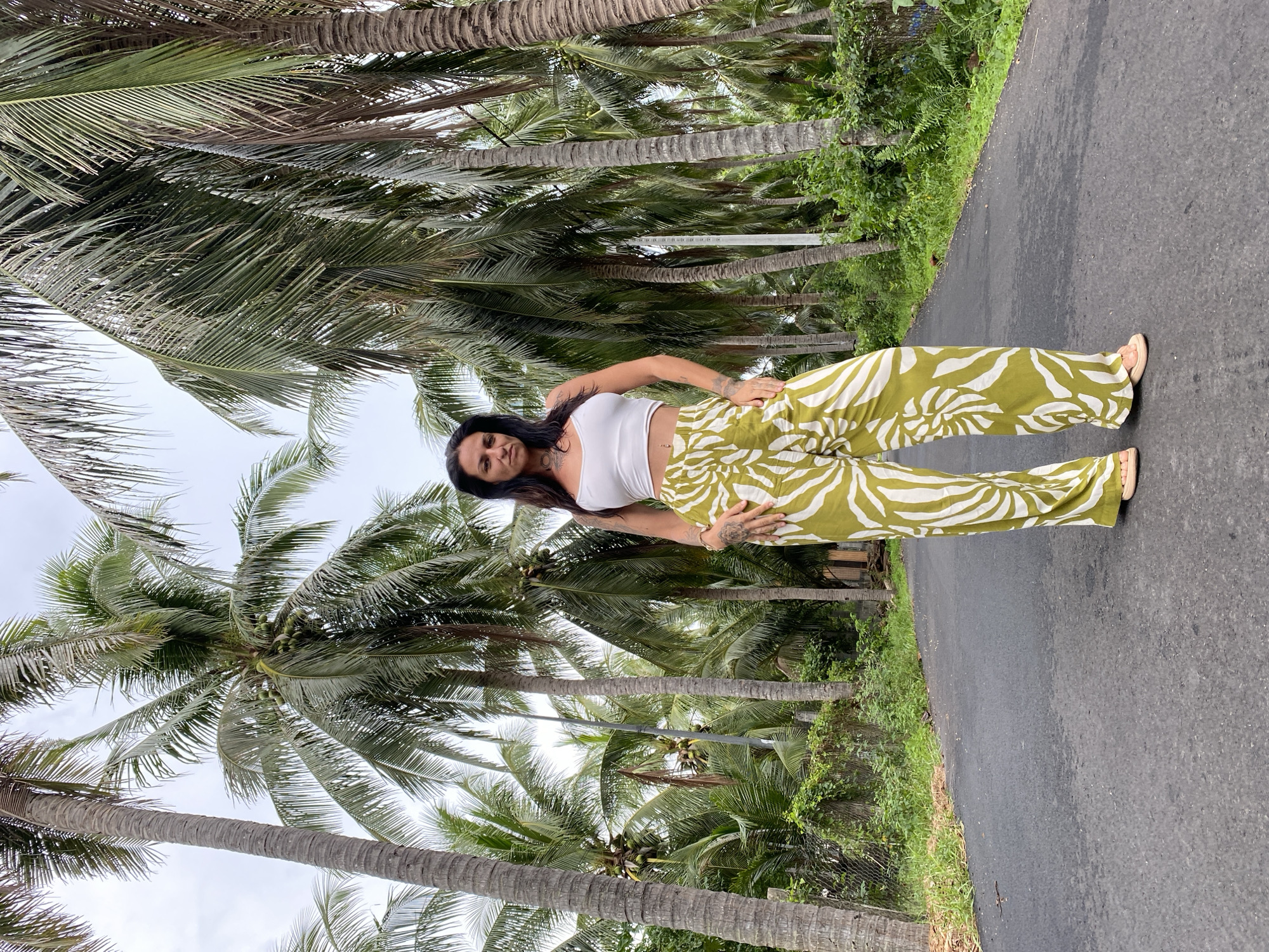 A woman stands on a paved road lined with lush green palm trees. She is wearing a white crop top and loose-fitting green and white patterned pants. The woman has dark hair and appears to be looking directly at the camera. The road is an asphalt surface, and on either side, there are dense clusters of tall palm trees with long, drooping fronds. The ground is a mix of asphalt and some scattered foliage. The sky is overcast. This scene is representative of the landscape found in Mũi Né, Vietnam, known for its extensive coconut palm groves.