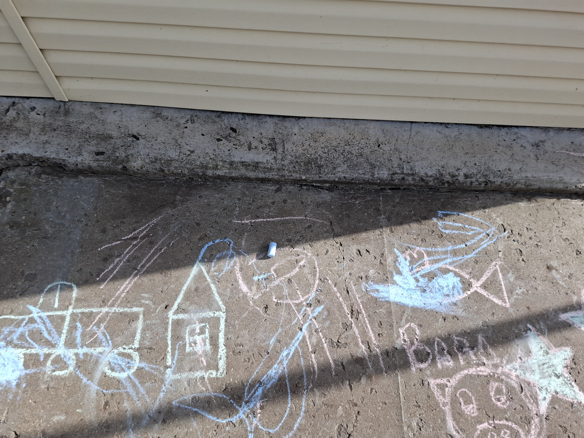 The image shows chalk drawings on a concrete surface, likely a sidewalk or foundation, in the city of Амзя, Russia. The drawings are rendered in various colors, including blue, pink, and white, and depict simple figures such as a house, a fish, a smiley face, and what appears to be the word "BABA" in block letters. A single piece of blue chalk lies on the concrete near the drawings. Above the concrete surface is a section of light-colored siding, possibly from a building. The lighting suggests it is daytime, with shadows casting across the ground. The concrete itself is weathered and shows some cracks and imperfections.