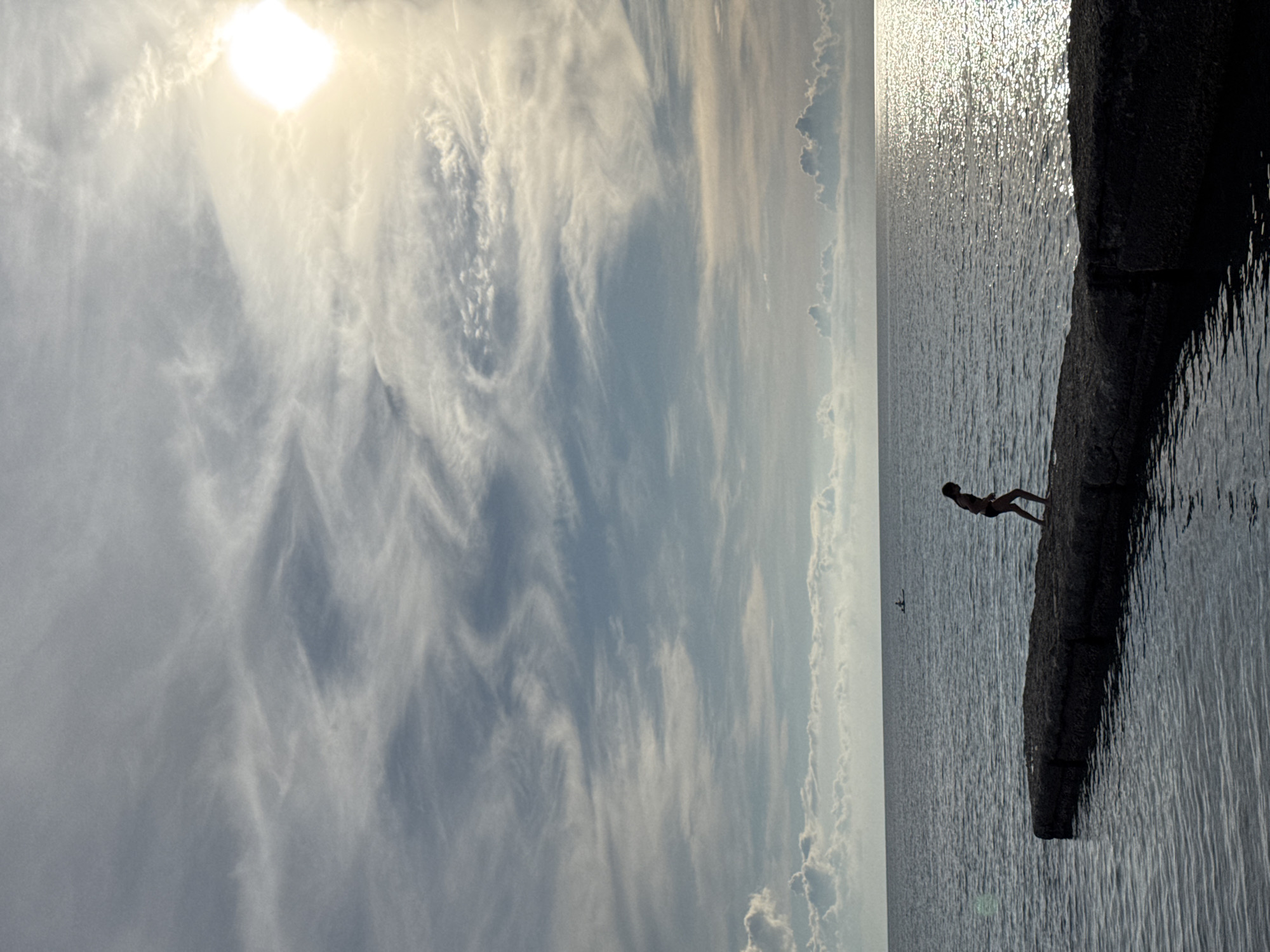 A solitary child in silhouette stands on a concrete pier extending into a calm, dark sea. The child is facing away from the viewer and appears to be walking or preparing to jump into the water. The sky above is dramatic, filled with wispy, white clouds that are illuminated by the bright, diffused light of the sun. The sun itself is partially obscured by clouds, creating a lens flare effect. In the distance, two figures on a small boat or kayak are visible on the water, adding a sense of scale. The scene suggests a serene, late afternoon or early evening ambiance, possibly at one of Sochi's many piers along the Black Sea coast. The water reflects the shimmering sunlight, creating a textured surface.