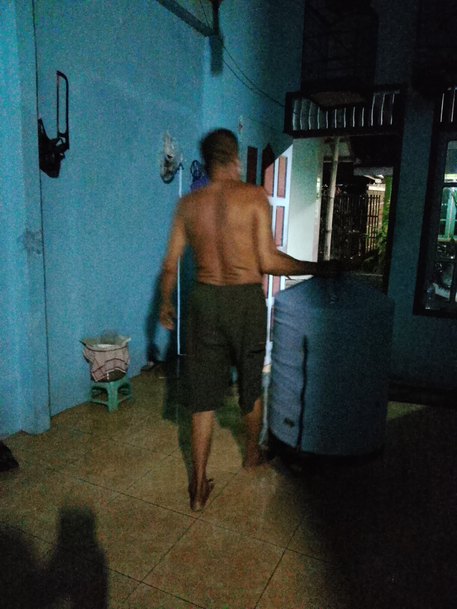The image depicts an indoor scene with a man seen from behind, walking away from the camera. The man is shirtless and wearing dark shorts. He appears to be in a dimly lit room with blue walls. To his right, there is a large, dark blue cylindrical object, possibly a water tank, and a doorway leading to an outdoor area with a fence. To his left, there is a small green stool with a bin containing a cloth. Shadows are cast on the floor, suggesting artificial lighting. The environment appears to be a residential setting within Kecamatan Pati, Indonesia, given the context provided.