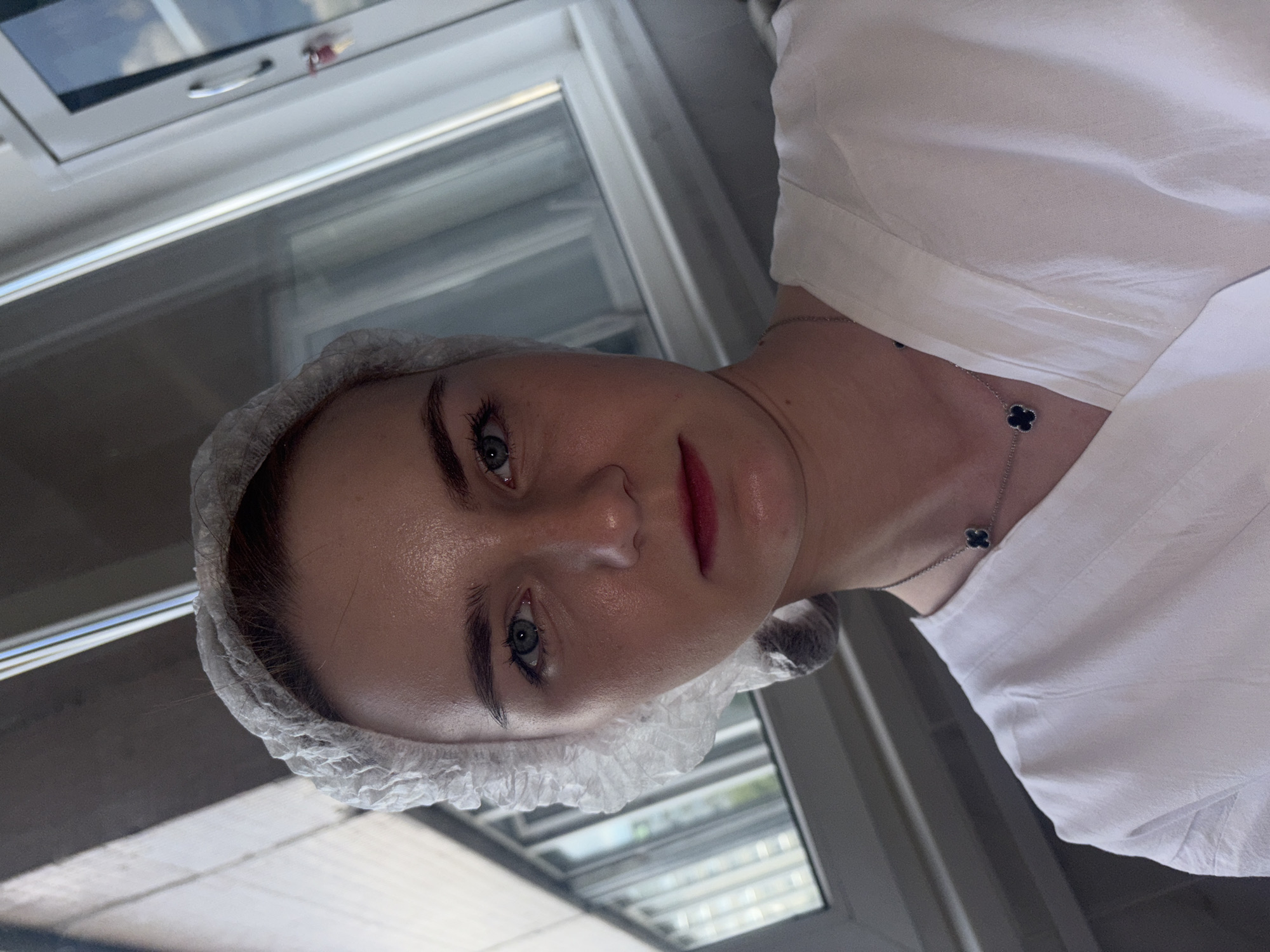 A young woman with blue eyes and blonde hair, wearing a white disposable hair net and a white V-neck top, is pictured indoors in front of a window. She is also wearing a silver necklace with a black clover pendant. The lighting suggests it is daytime. The background includes reflections in the window and a window frame with a handle.