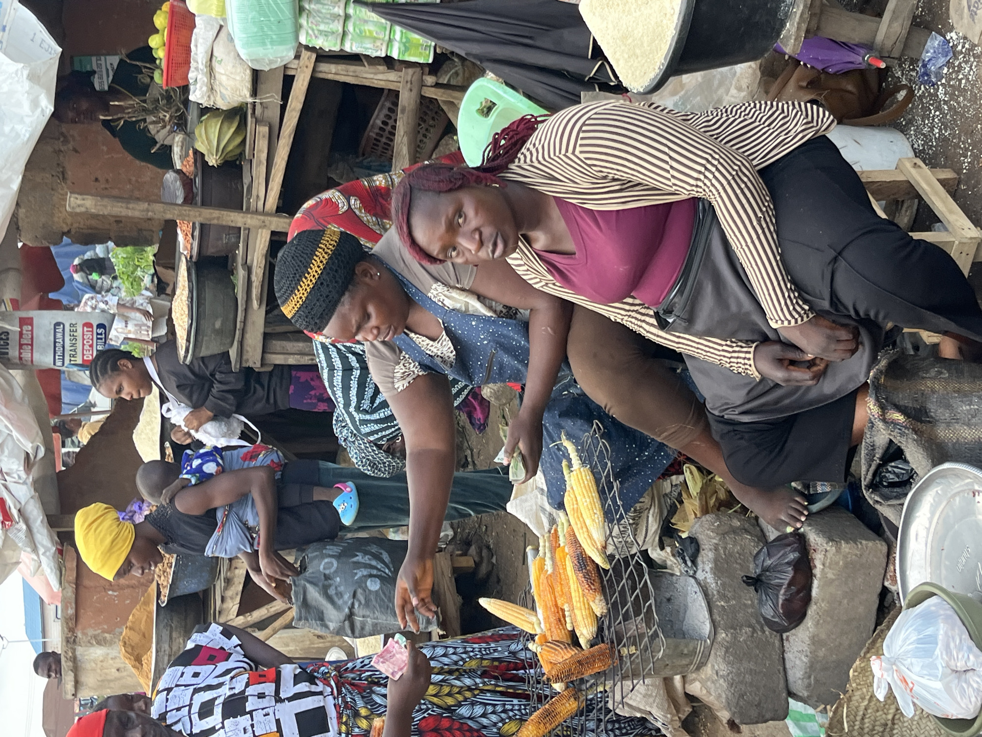 This media file captures a lively scene at an outdoor market in Bukuru, Nigeria, likely during daylight hours given the ambient lighting. The focus is on several individuals, predominantly women, engaged in commerce. One woman, wearing a striped long-sleeved top and dark trousers, leans forward, her expression seemingly focused and perhaps a bit weary. Beside her, another woman, adorned with a black and gold patterned head covering and a blue patterned dress, is also leaning in, her attention directed downwards.
The setting is a bustling market stall, characterized by rustic wooden structures and an array of goods. Visible objects include piles of corn on a grill, bags of produce or grains, and various containers. The ground is somewhat cluttered with scattered materials, suggesting a well-used commercial space. A young child, partially visible and carried by an adult, adds to the sense of community and daily life. Another individual in the foreground is partially visible, wearing a colorful patterned garment.
The activity appears to be the sale of goods, with hands reaching out, possibly exchanging money or examining products. The presence of cash being held by one person suggests a transaction is in progress. The overall atmosphere seems to be one of energetic marketplace activity, a common sight in many Nigerian locales. There are no immediately discernible weather cues, but the open-air setting implies it is not actively raining. The location cues are limited to the context provided, but the visual elements are consistent with a typical West African market.