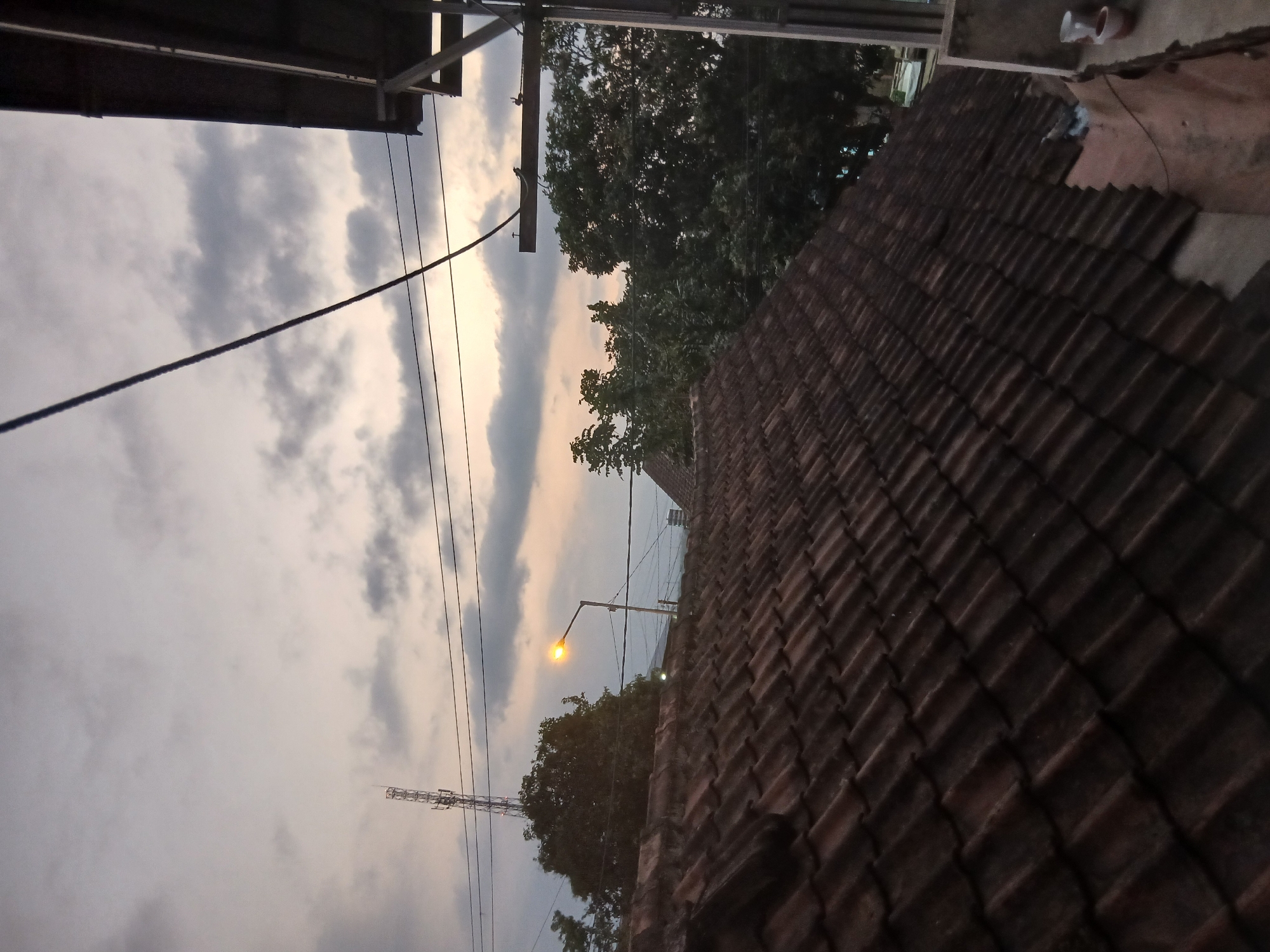 The image shows a view from a rooftop in Kademangan Kulon, Indonesia, at dusk. The foreground is dominated by a terracotta tiled roof. Beyond the roof, there are trees and a street lamp with an illuminated yellow bulb. In the distance, a cell tower is visible against a cloudy sky. Overhead, power lines crisscross the scene. The overall impression is of a residential or semi-urban environment during twilight.