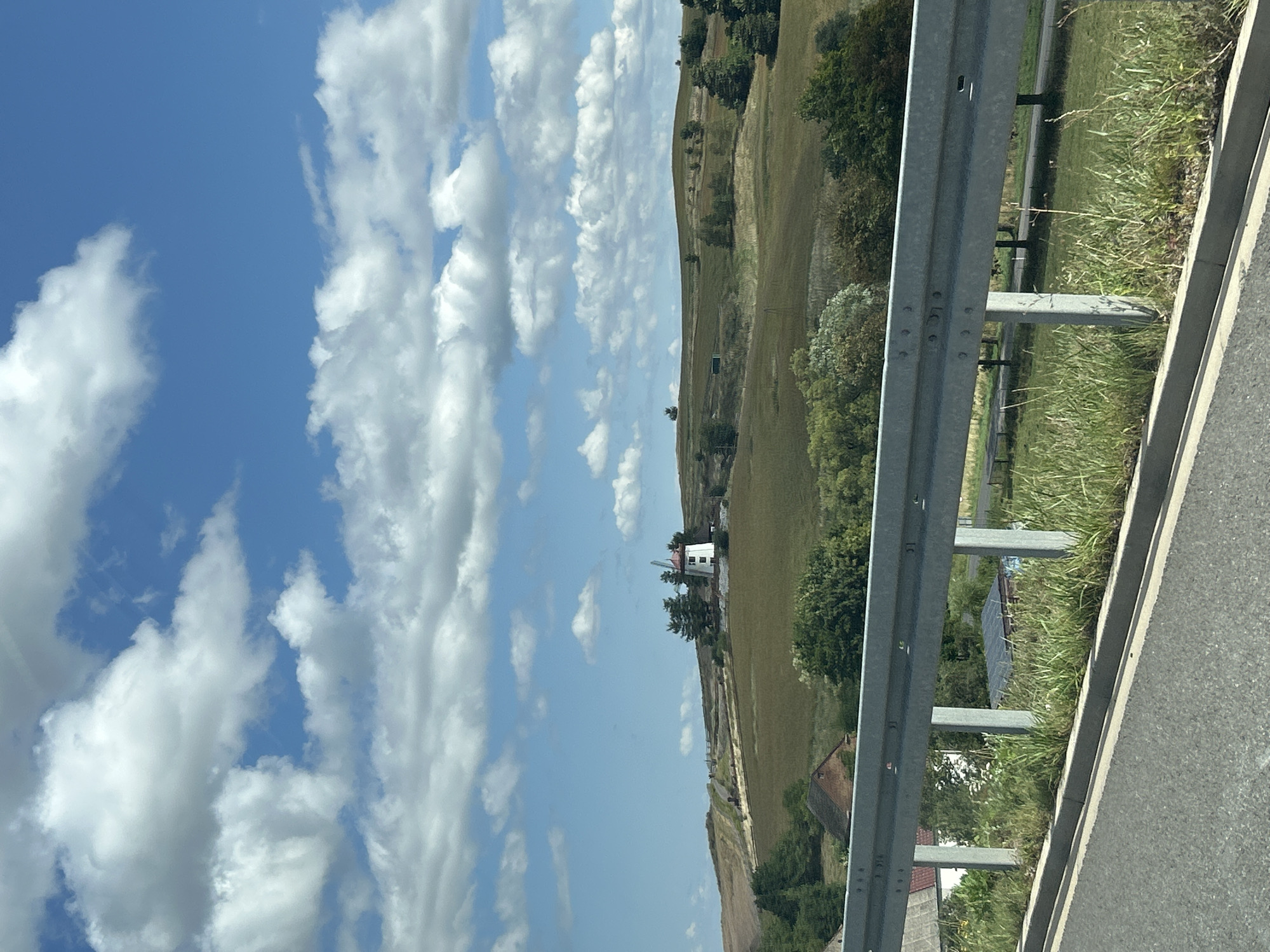 The image depicts a picturesque landscape with a prominent windmill on a rolling green hill under a bright blue sky dotted with fluffy white clouds. The windmill is white with a red roof and appears to have four sails. The surrounding terrain is characterized by grassy slopes, scattered trees, and a few buildings nestled in the lower parts of the hill. In the foreground, a metallic guardrail runs along the side of an asphalt road, with a strip of overgrown grass and vegetation separating the road from the embankment. The scene suggests a rural setting in Gostków, Poland, possibly a viewpoint or a roadside attraction. There are no people or animals visible in the image.
