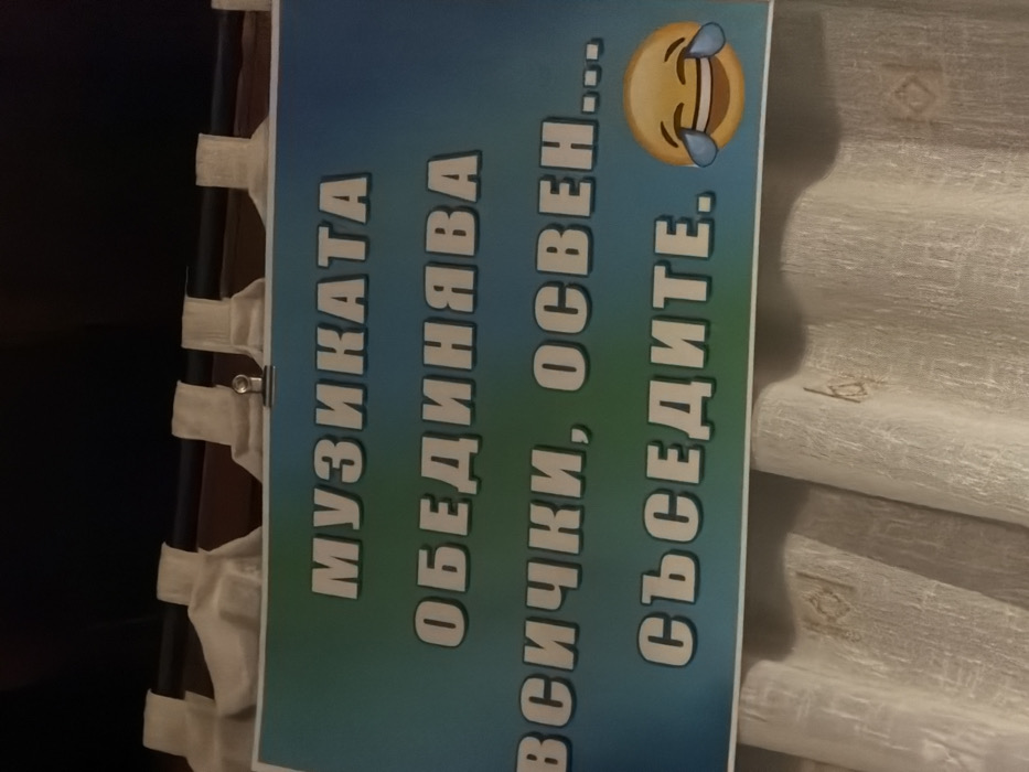 The image displays a rectangular sign in Turiya, Bulgaria, hung indoors above light-colored curtains. The sign features a blue-to-green gradient background with white Cyrillic text that reads: "МУЗИКАТА ОБЕДИНЯВА ВСИЧКИ, ОСВЕН... СЪСЕДИТЕ." (Music unites everyone, except... the neighbors.) A yellow "face with tears of joy" emoji is positioned to the right of the last word. The sign is attached to a dark curtain rod by at least one silver metal clip visible at the top center. The curtains below the sign exhibit vertical folds and a subtle patterned texture. A dark, indistinct area, possibly a wall or ceiling, is visible above the curtain rod. No people are present in the scene.