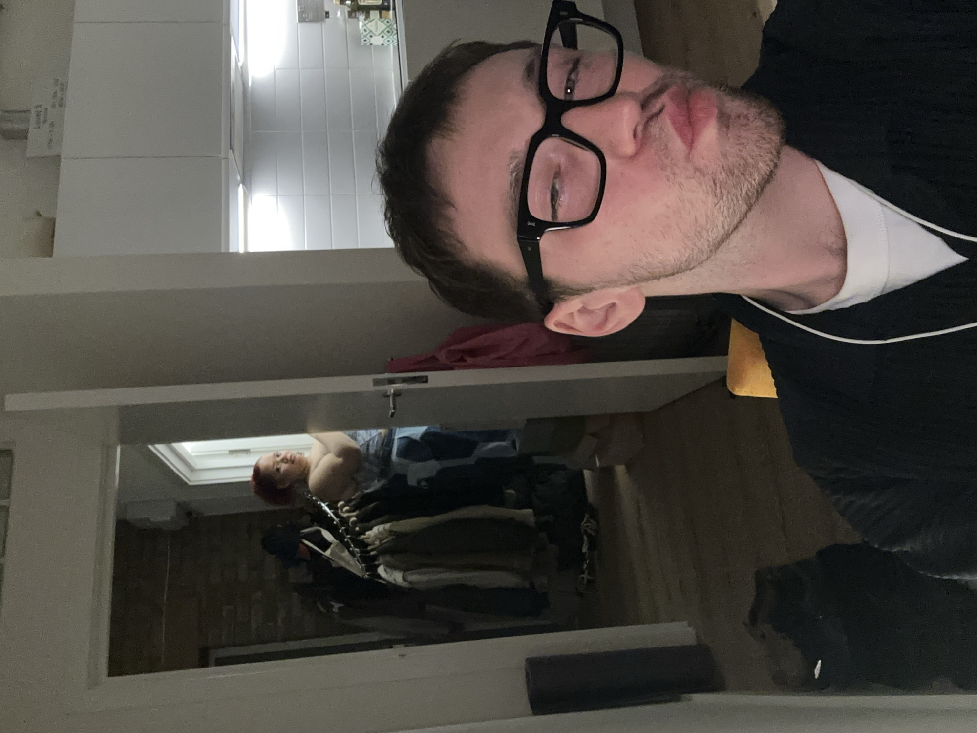 The photograph shows a man with glasses and stubble, wearing a white t-shirt under a black top, partially visible in the foreground. In the background, seen through an open wardrobe or doorway, there is a person with red hair lying down. The wardrobe contains clothes on hangers. The location seems to be an indoor setting. Based on the style of architecture and interior design, the image could potentially be set in London, United Kingdom, although definitive confirmation of location based on the image is not possible.