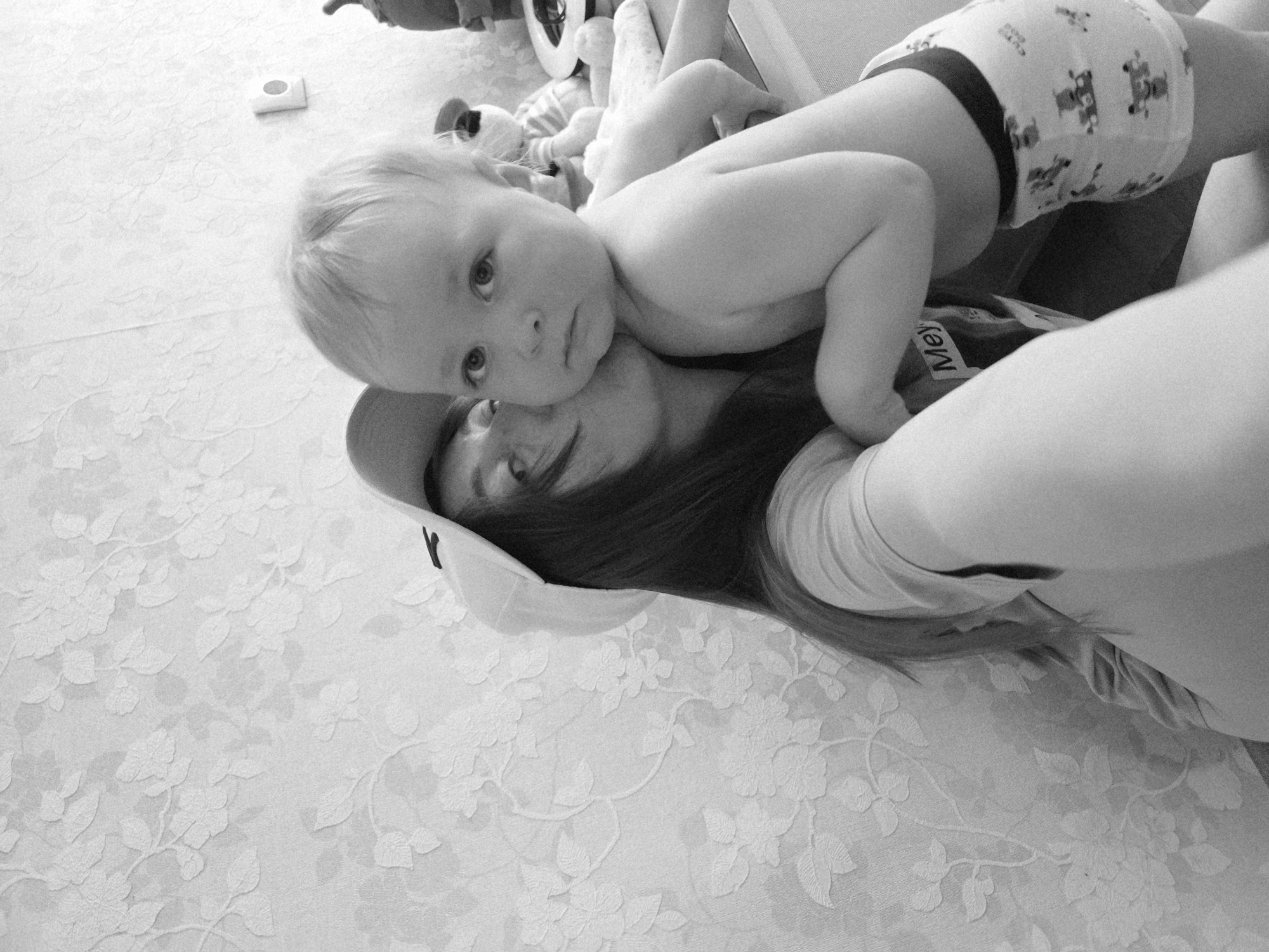 A black and white photograph captures a mother lying on a white floral-patterned surface, possibly a bed, with her baby lying on top of her chest. The baby, wearing patterned underpants, faces forward with a somewhat contemplative expression. The mother, wearing a cap, looks up at the camera. There's a partial view of other objects and toys blurred in the background. The image has a slightly upward angle. Based on the provided context of Chaikovsky, Russia, it can be inferred that this intimate moment is captured in a domestic environment within this city.