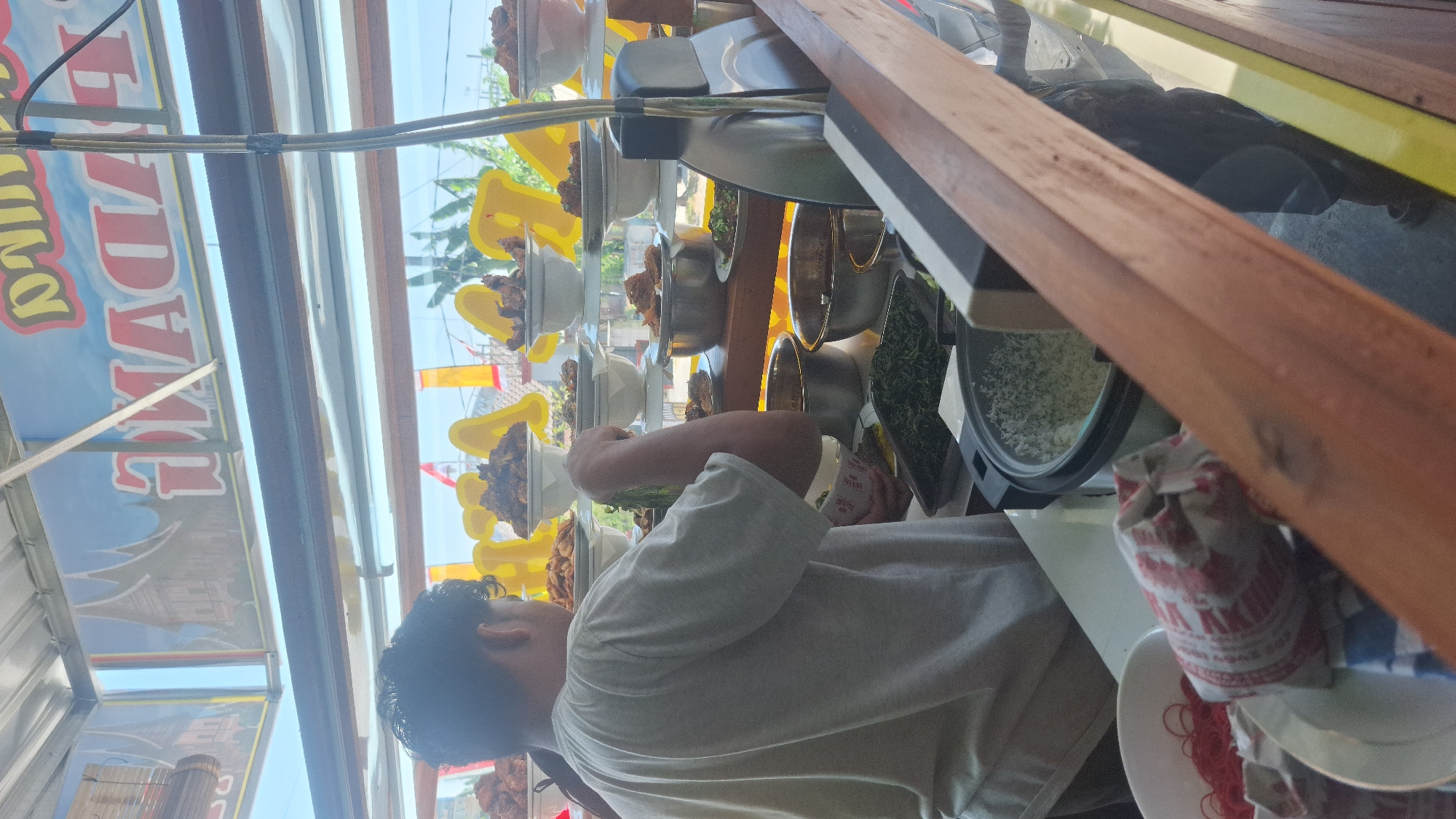 The image depicts a person, likely a vendor, serving food at an outdoor food stall in Widodaren, Indonesia. The vendor, seen from behind, wears a grey t-shirt and is in the process of handling a bunch of green leafy vegetables. The stall displays an array of food items, predominantly served in white bowls and platters, including what appears to be fried chicken or other meats. A rice cooker is visible, filled with white rice. The background shows a blue sky with hints of green foliage, suggesting an outdoor setting. The stall is decorated with banners, one of which displays text and images, possibly indicating the name of the establishment or promotional material. The overall scene is one of a typical Indonesian street food vendor preparing and serving meals.