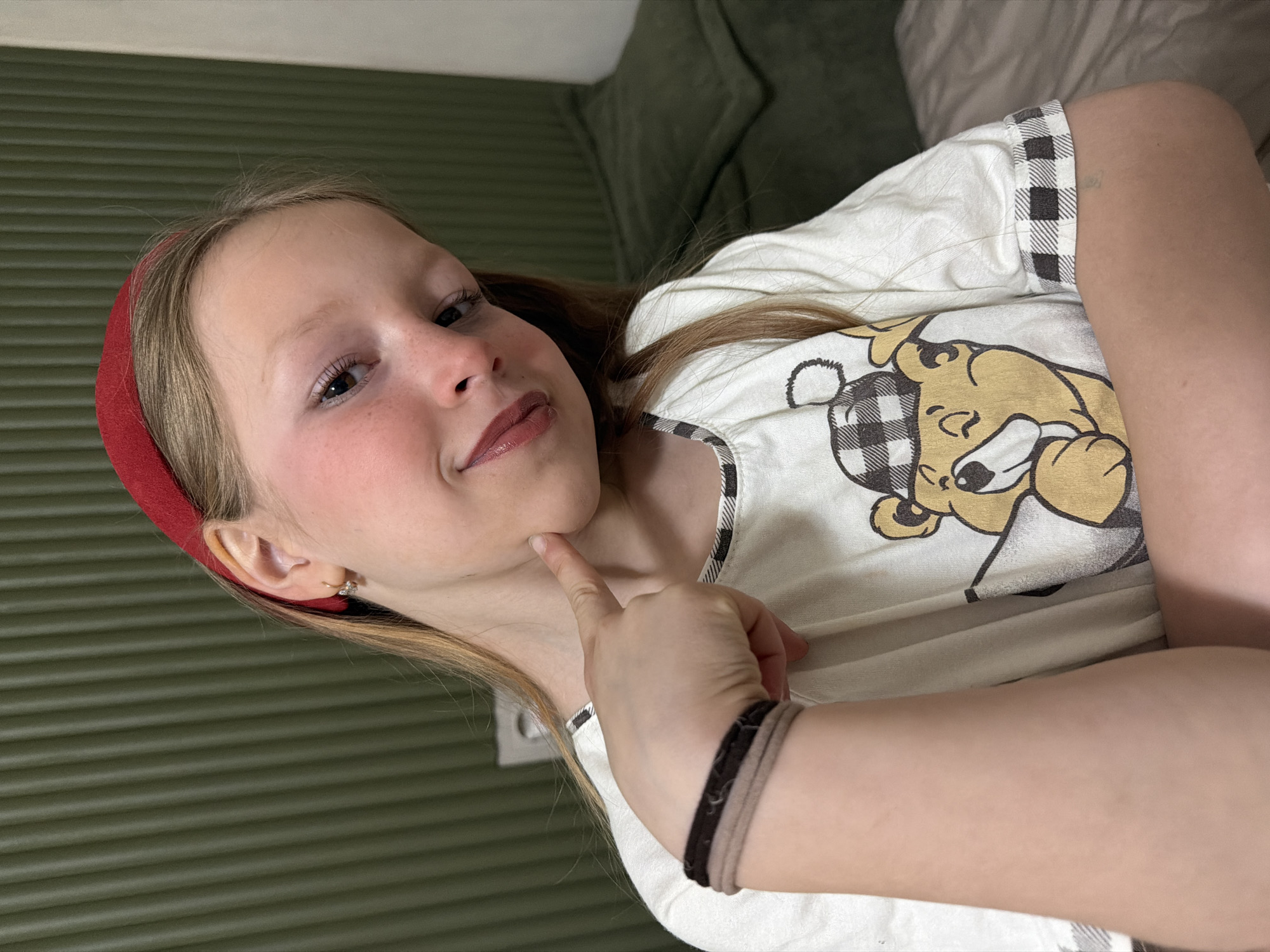 The image features a young girl, likely between 8 and 12 years old, posing indoors in Dubovaya, Russia. She has fair skin, light brown hair, and dark eyes, enhanced with light pink lipstick or lip gloss. A red headband adorns her hair, and she wears a small hoop earring in her right ear. She is dressed in a white short-sleeved top featuring a cartoon character on the chest – a light brown bear wearing a black and white plaid shirt. On her right wrist, she has two elastic hair ties or simple bracelets, one dark brown and one beige.
The girl is looking directly at the camera with a gentle, slightly playful smile, her right index finger softly touching her chin in a thoughtful or coy gesture.
The background consists of a dark olive-green wall with horizontal ribbed or slatted paneling. In the upper right corner, a portion of a dark green upholstered sofa or armchair is visible. The lighting is soft, indicating an indoor setting. The image is oriented diagonally, with the girl's head tilted towards the upper left. There is no visible text in the image.