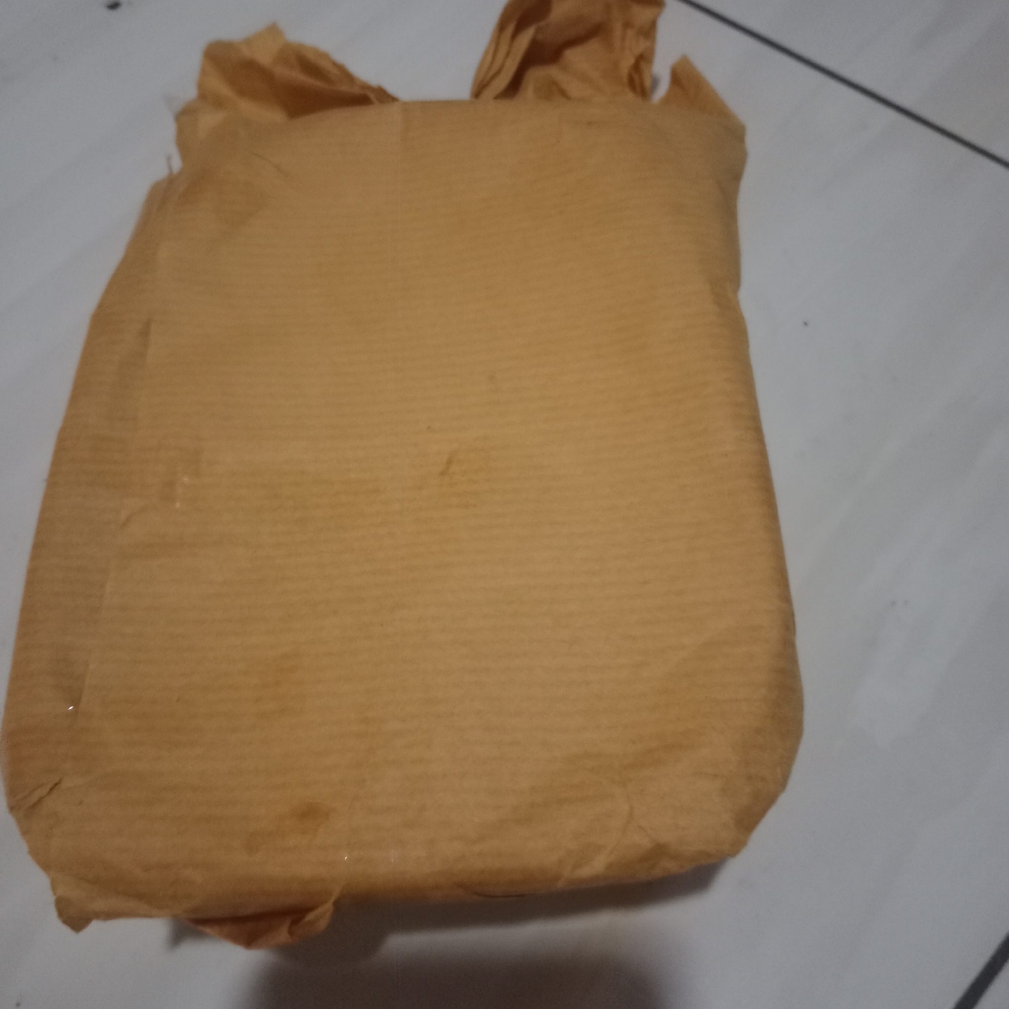 A brown, rectangular package is positioned on a light-colored, tiled surface. The package appears to be wrapped in textured brown paper, with some areas showing slight creases and wrinkles. The background consists of a tiled floor, with thin dark lines indicating grout lines. The lighting in the scene is dim, casting a subtle shadow to the right of the package. The image was taken in Tegowanu Kulon, Indonesia.